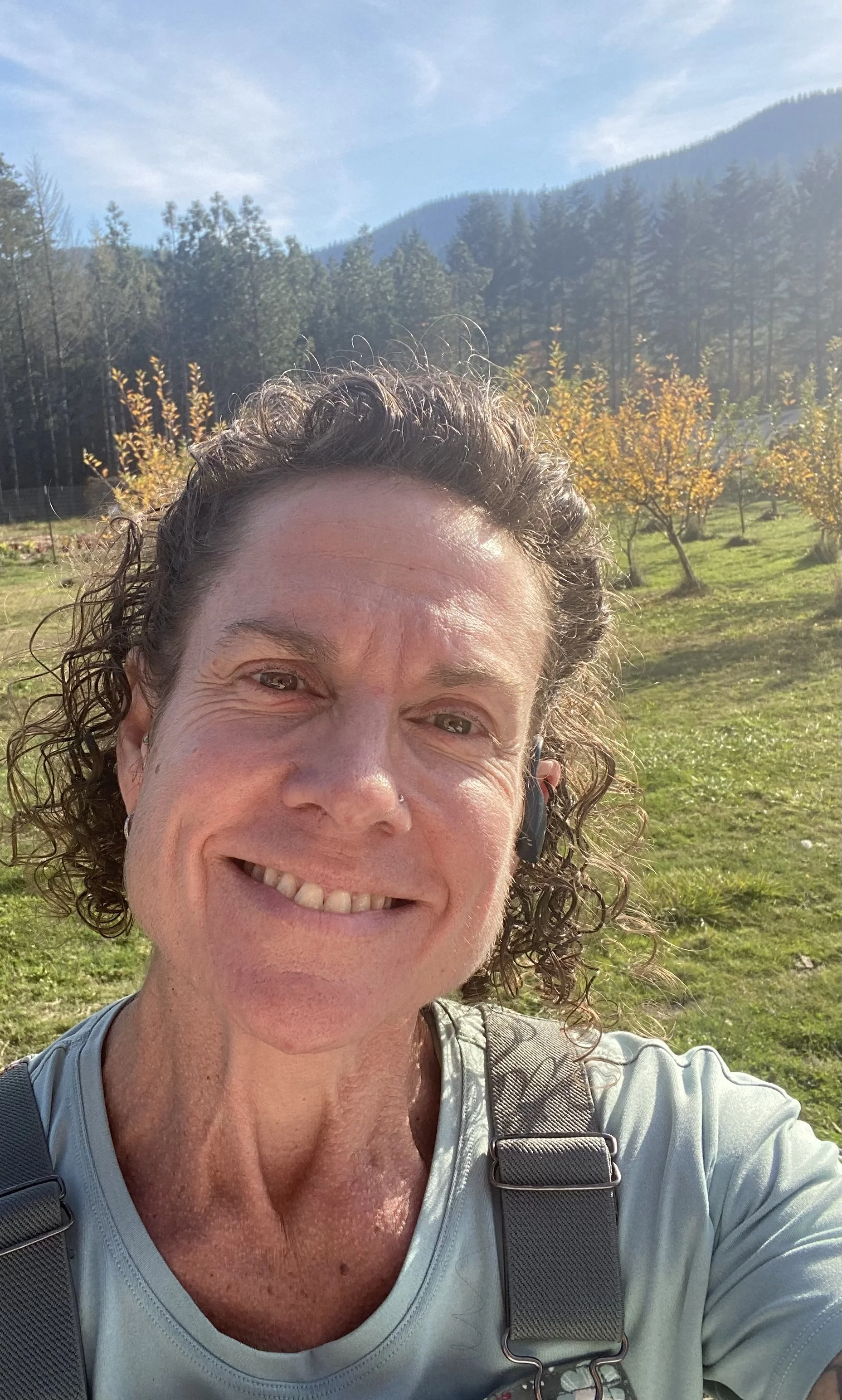 A smiling woman taking a selfie outdoors with a background of trees and mountains on a sunny day.