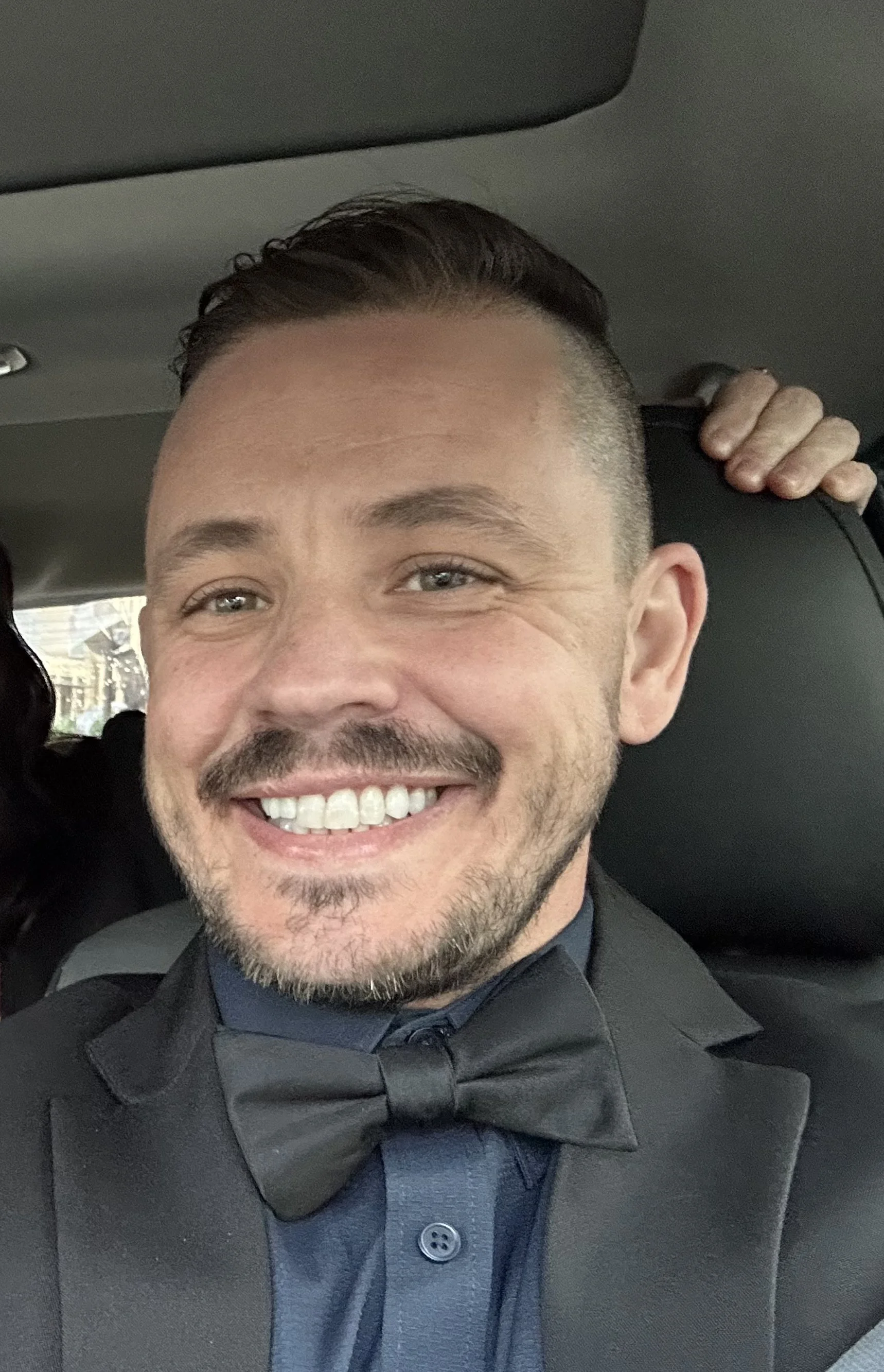 A man smiling in a tuxedo, sitting inside a vehicle.