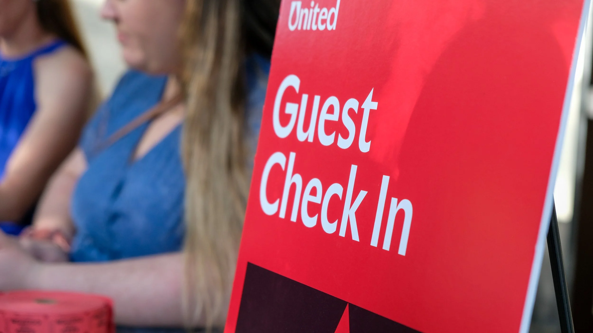 A red sign with white text reading 'Guest Check-In' placed on a stand at an event. Two women are in the background, blurred, sitting at a table.