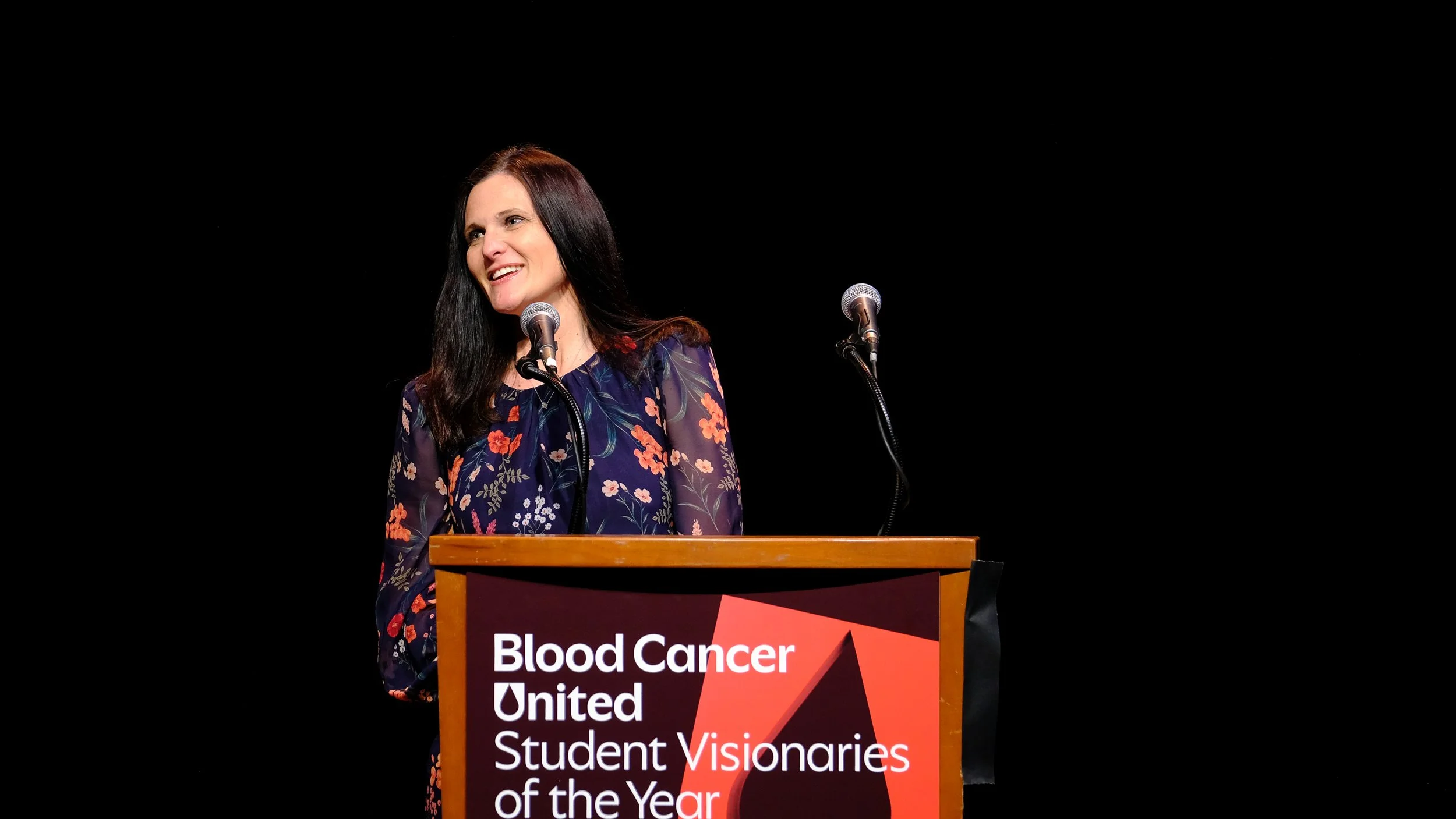 A woman with long dark hair smiling and speaking at a podium with two microphones during a Blood Cancer United event, with a black background.