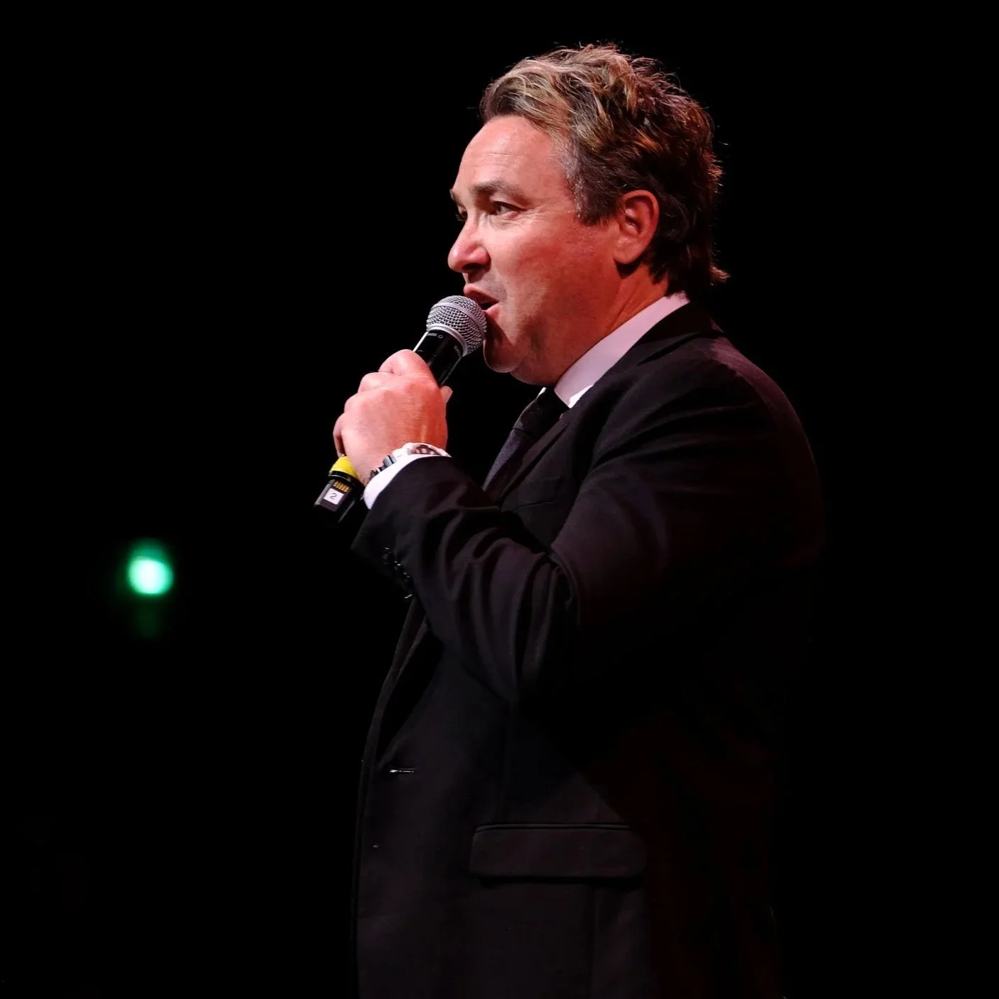 A man in a suit holding a microphone, speaking or singing on stage against a black background.
