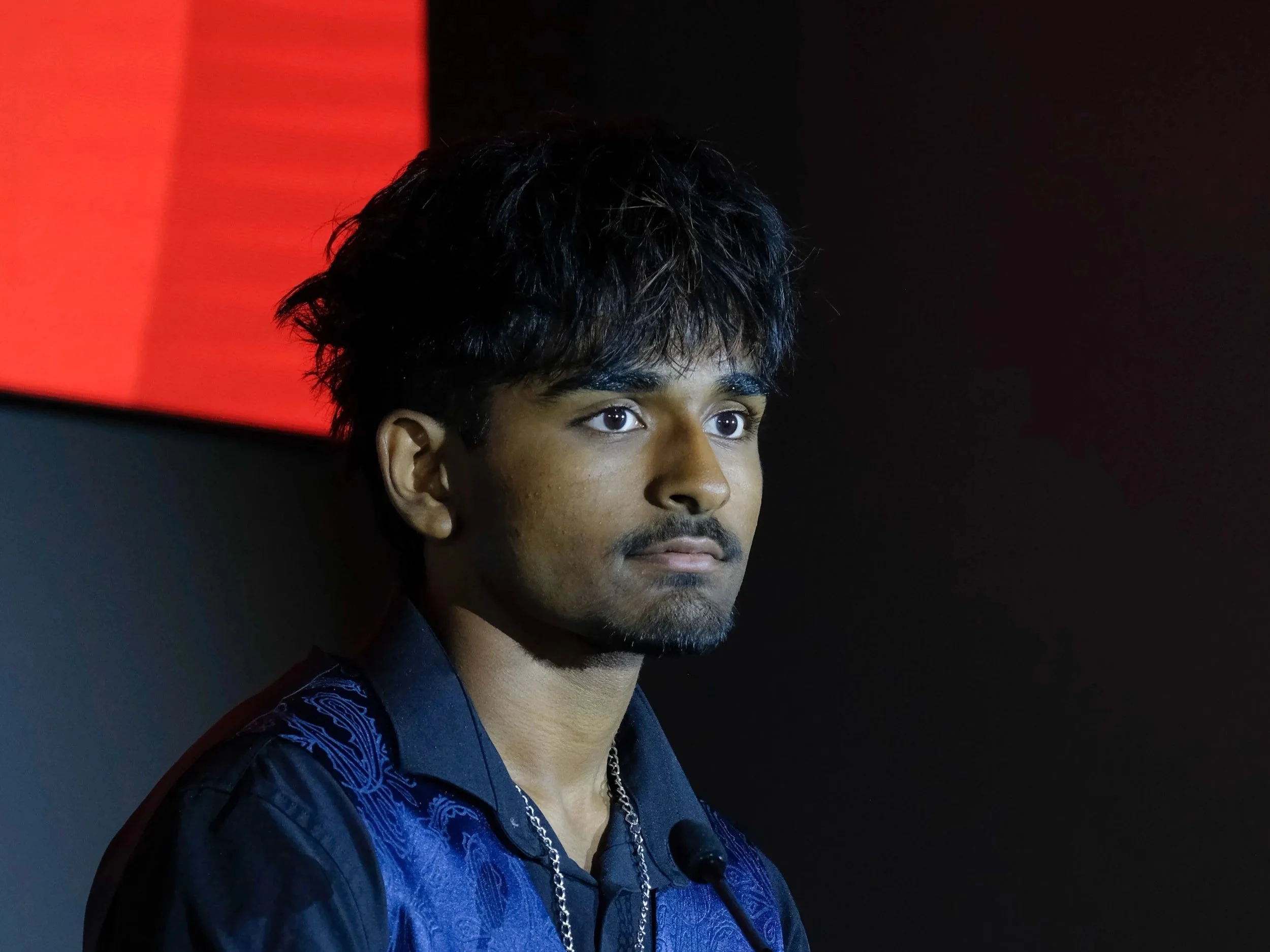 A young man with dark, curly hair and a beard, wearing a dark blue shirt with a subtle pattern and a silver chain, appears to be at an event with a red and black background.