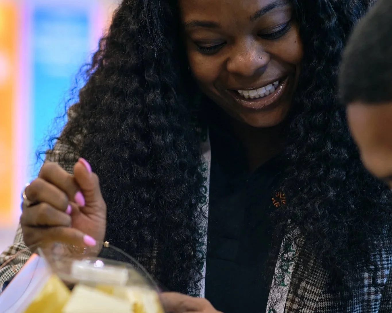 A smiling woman with long, curly black hair and a nose piercing, engaged in conversation with a young boy. She is wearing a checkered jacket and has pink nails. There is a plate of lemon slices in the foreground.