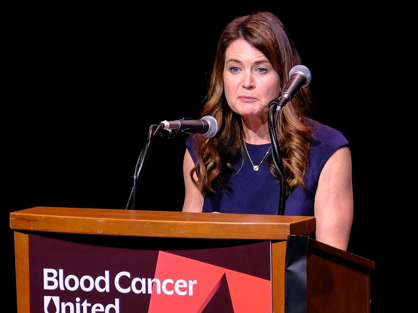 A woman with long brown hair speaking at a podium with a sign that reads 'Blood Cancer United' behind her. She is wearing a dark blue sleeveless dress and a necklace, and is speaking into two microphones.