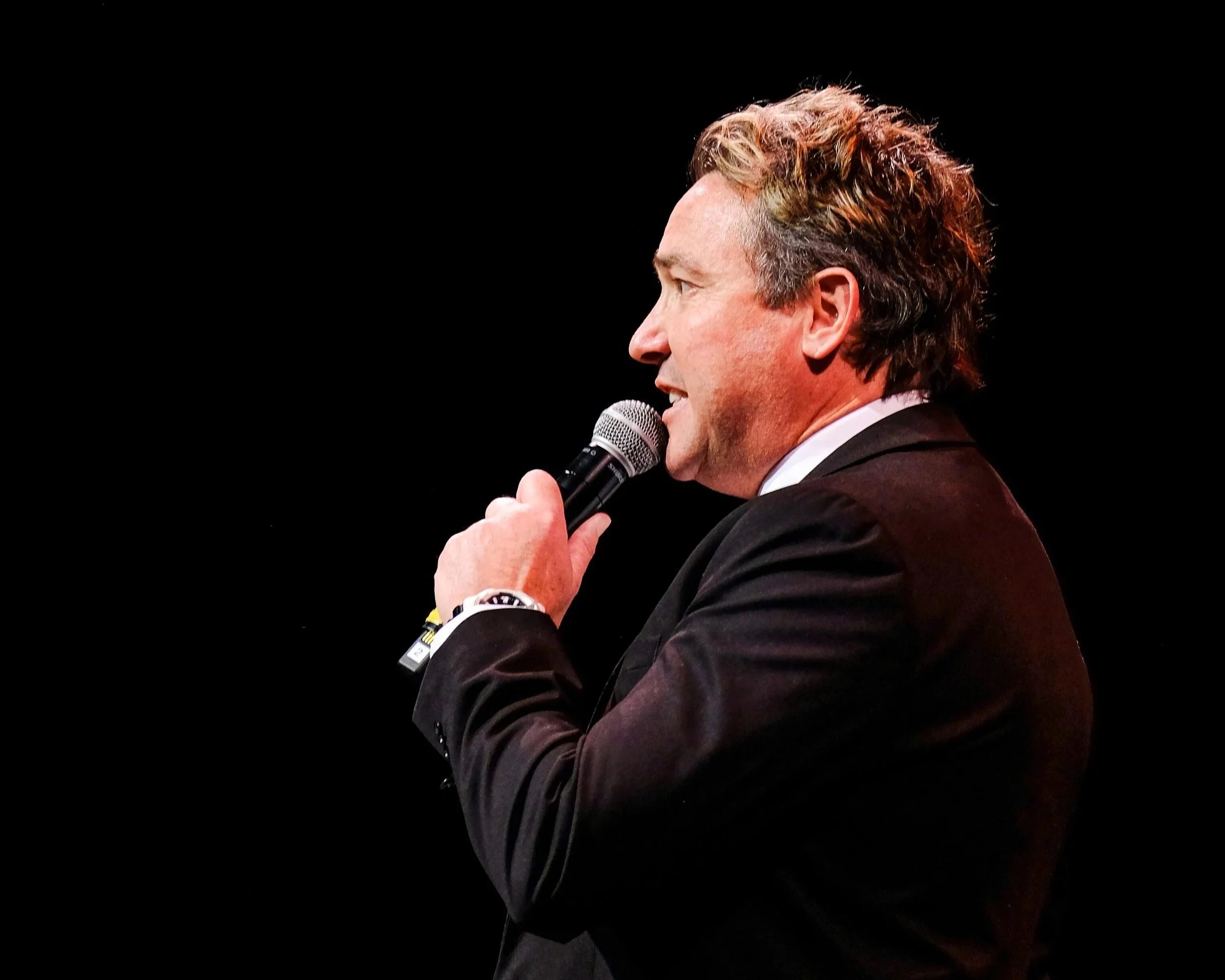 A man holding a microphone, dressed in a dark suit, speaking on stage with a black background and distant city lights.