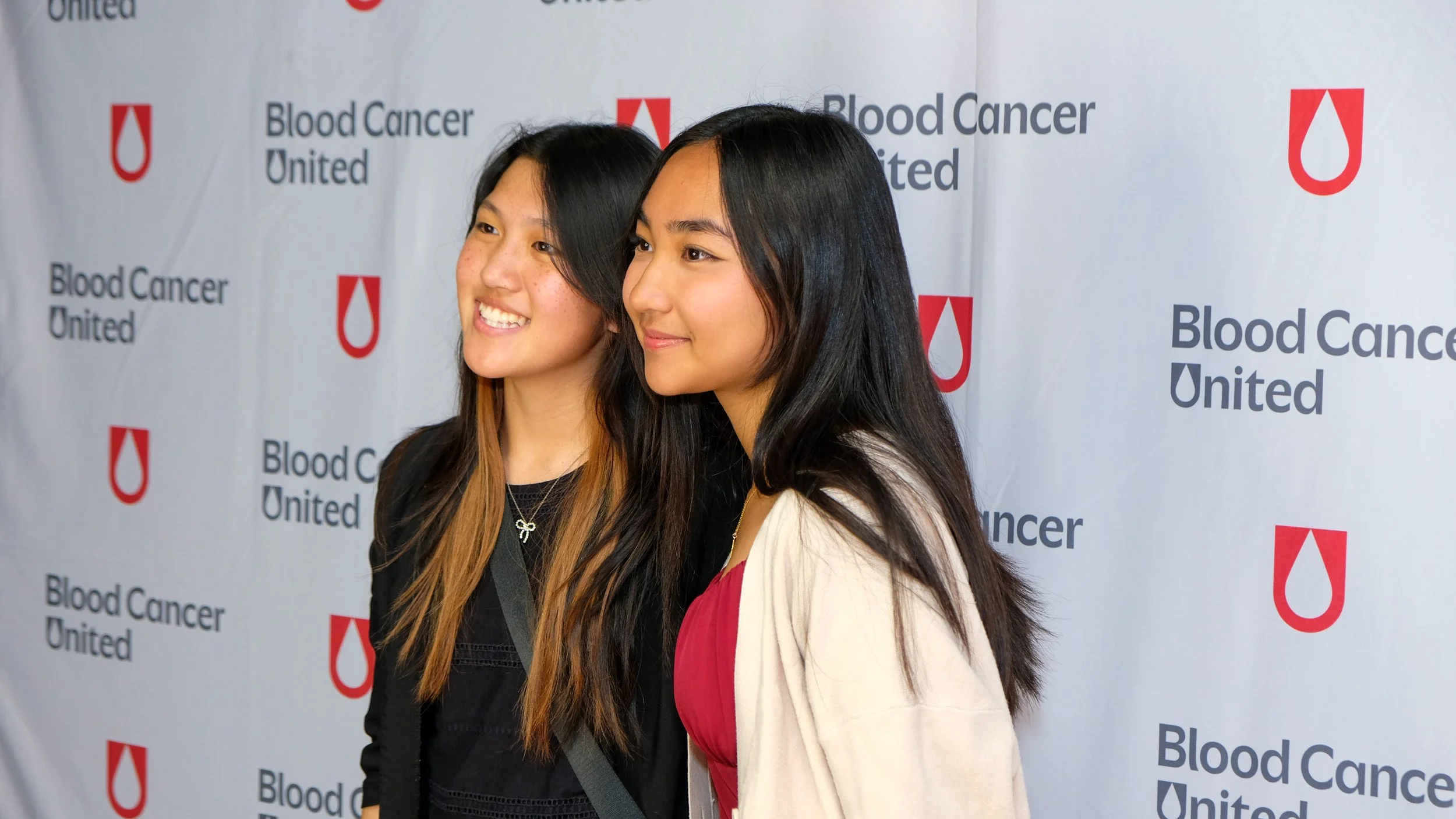 Two women standing together at a Blood Cancer United event, smiling, with a backdrop displaying the organization's logo and name.