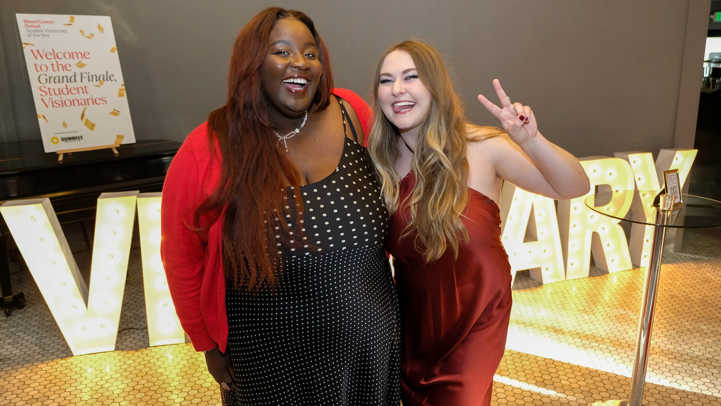 Two women smiling and posing for a photo at an event, with illuminated letters spelling 'VY' in the background and a welcome sign for students and visionaries to the Grand Finale.