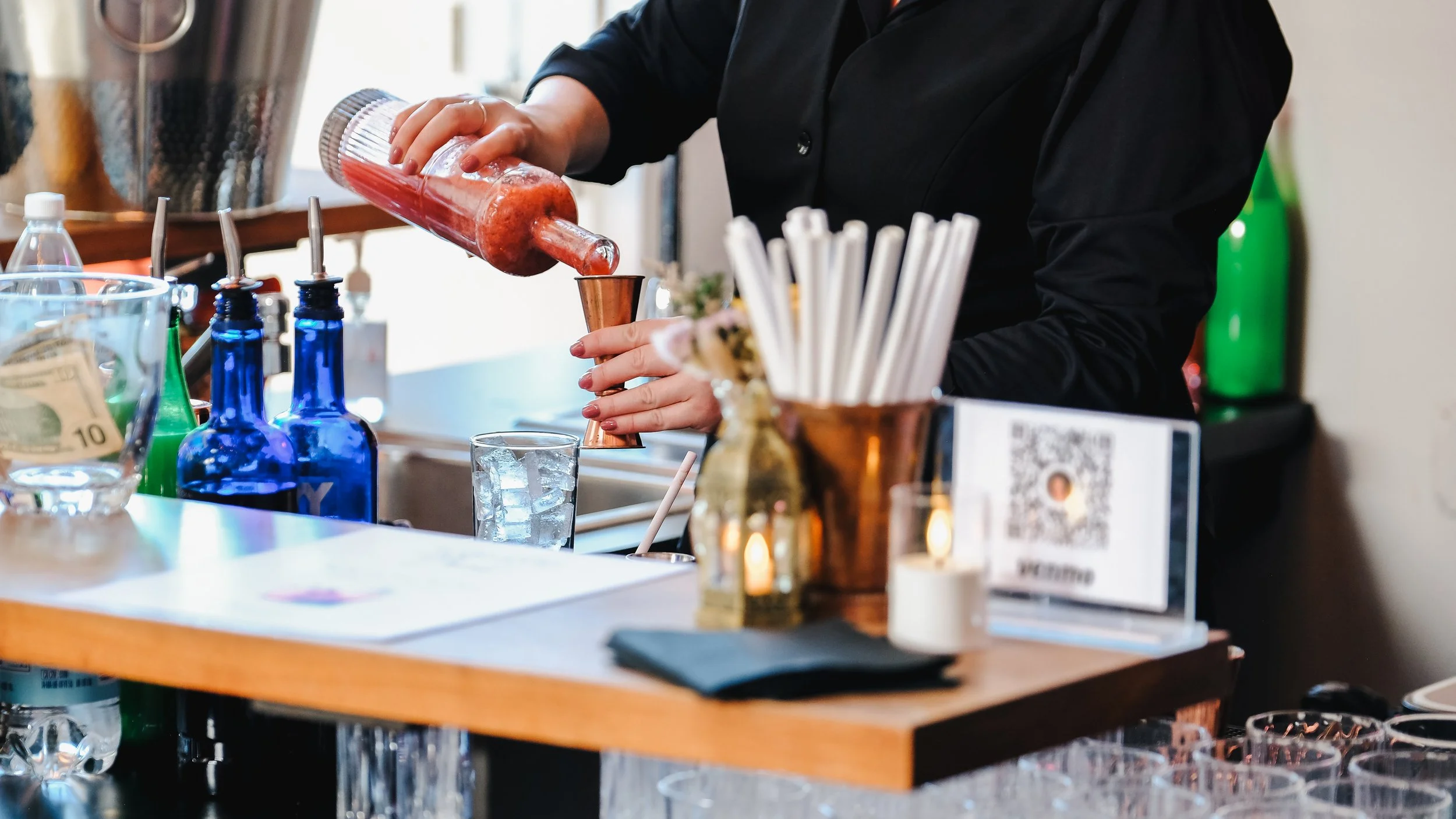 A bartender in black attire pouring a red cocktail from a shaker into a copper cup at a bar with various bottles and bar tools visible.