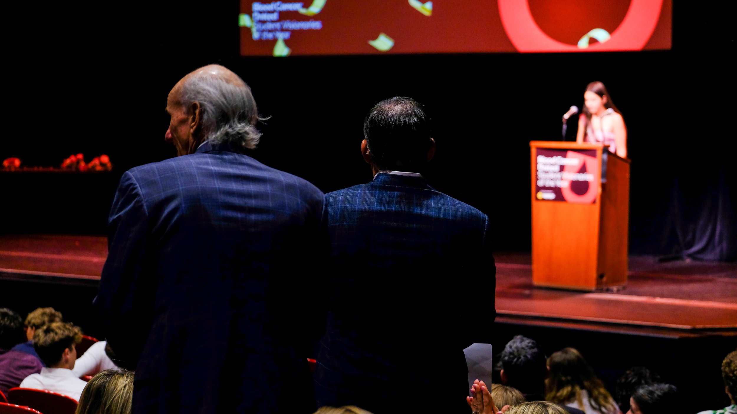 At a conference podium with a signer on stage, audience members seated, two men in suits standing in the foreground, large screen displaying a red graphic element and text in the background.