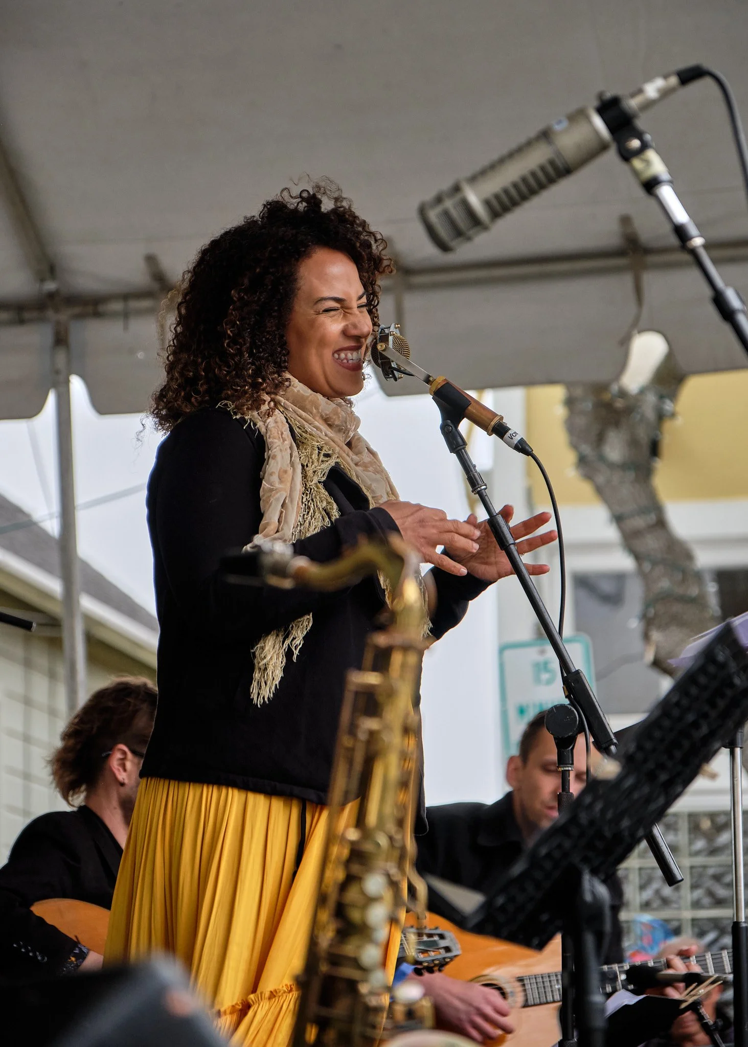 A woman with curly hair, wearing a beige scarf and black top, is singing passionately into a microphone while playing a wind instrument. Other musicians are partially visible around her, including a person playing a guitar and others with musical instruments, under a tent at an outdoor event.