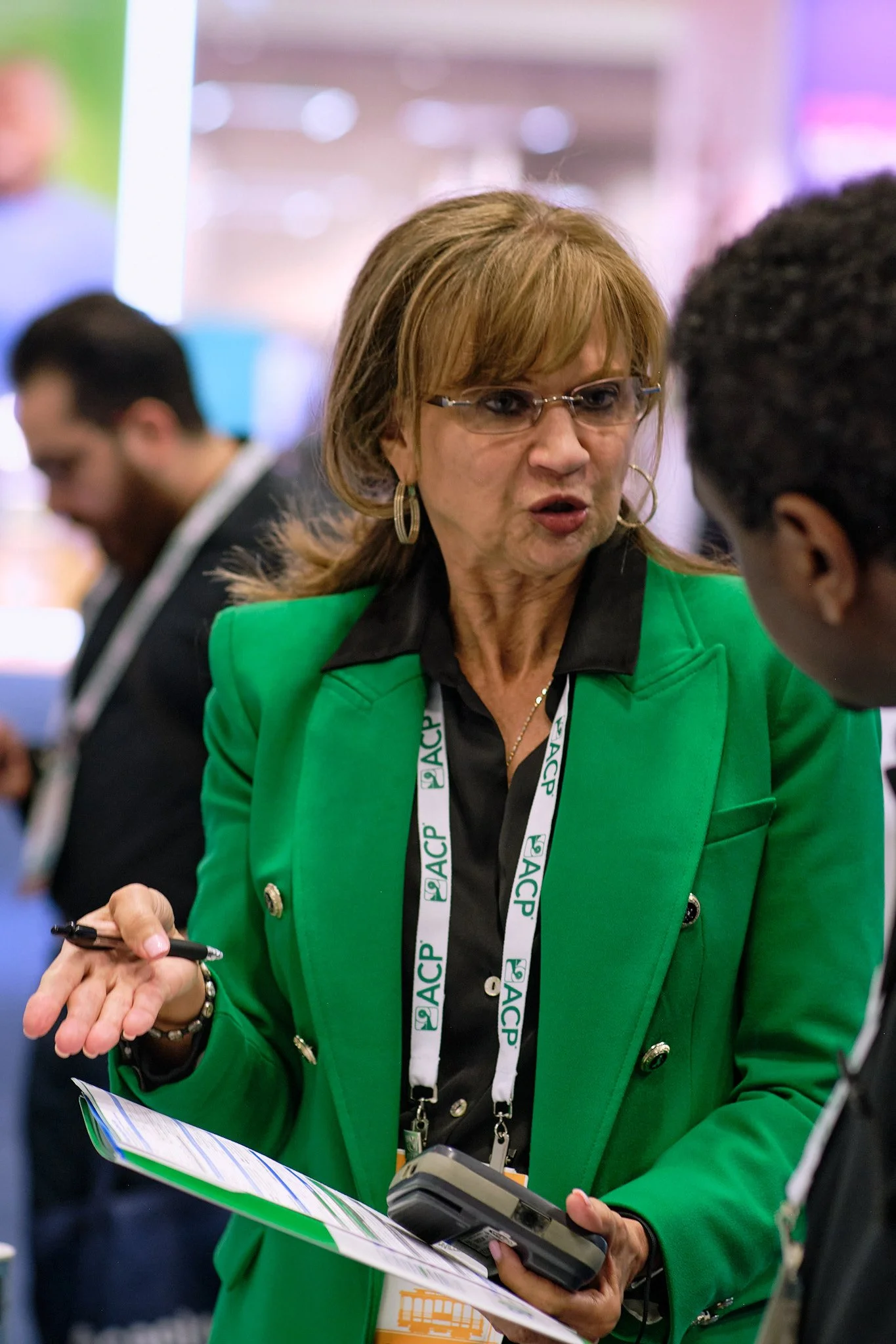 A woman with glasses and earrings wearing a bright green jacket is engaged in conversation with a person holding a device. She has a lanyard around her neck with the letters ASCIP. There is a man in the background wearing a dark suit.