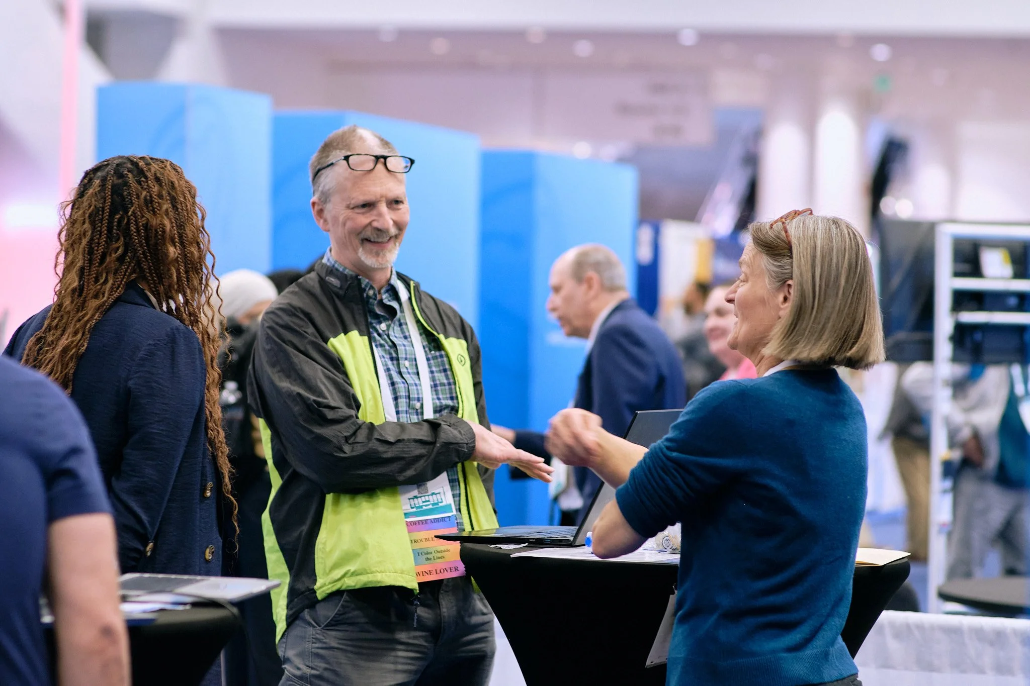 People at a conference or event, engaging in conversation, with one person shaking hands across a table, in a bright indoor setting.