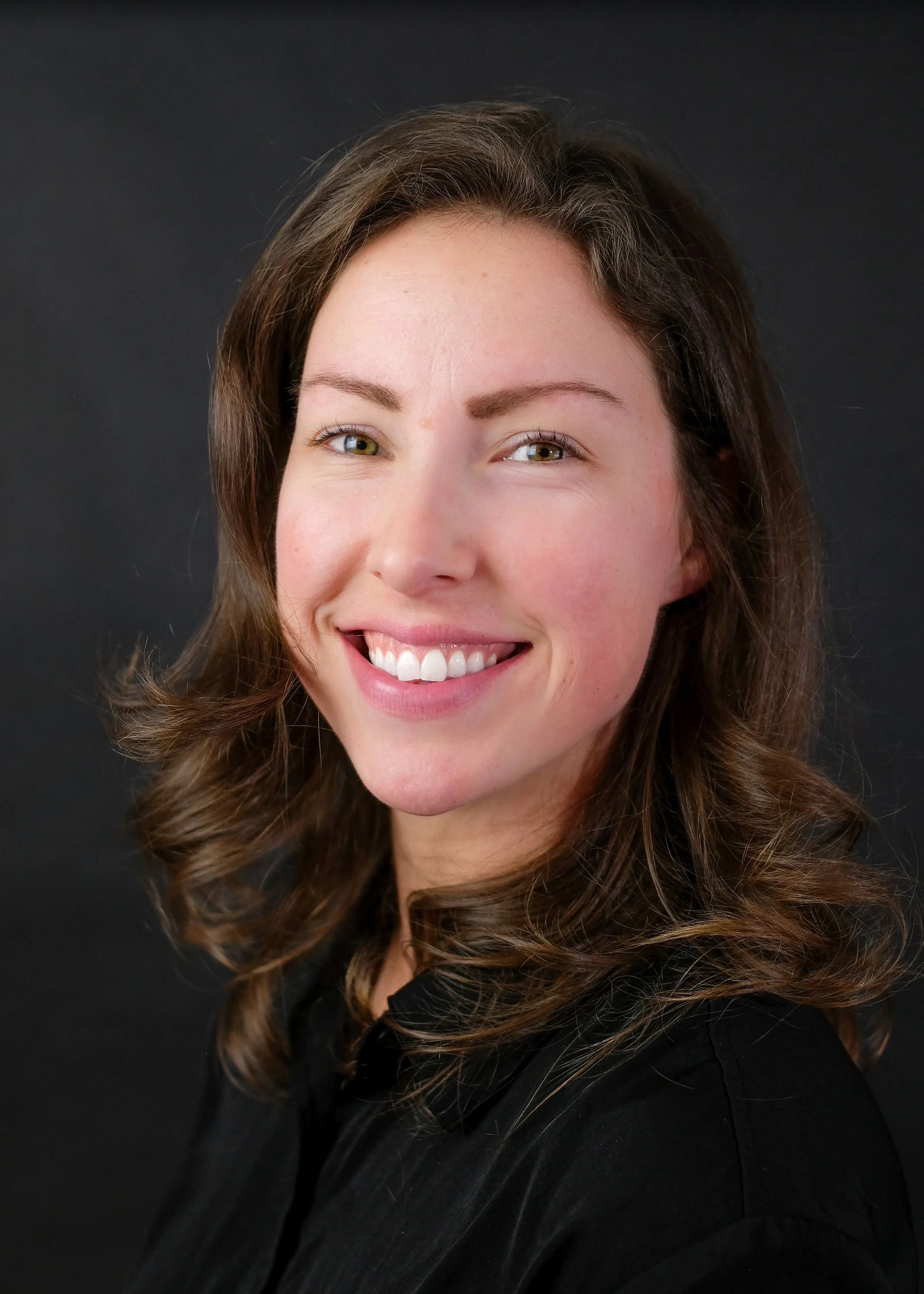 Close-up portrait of a woman with brown curly hair, smiling, wearing a black top, against a dark background.
