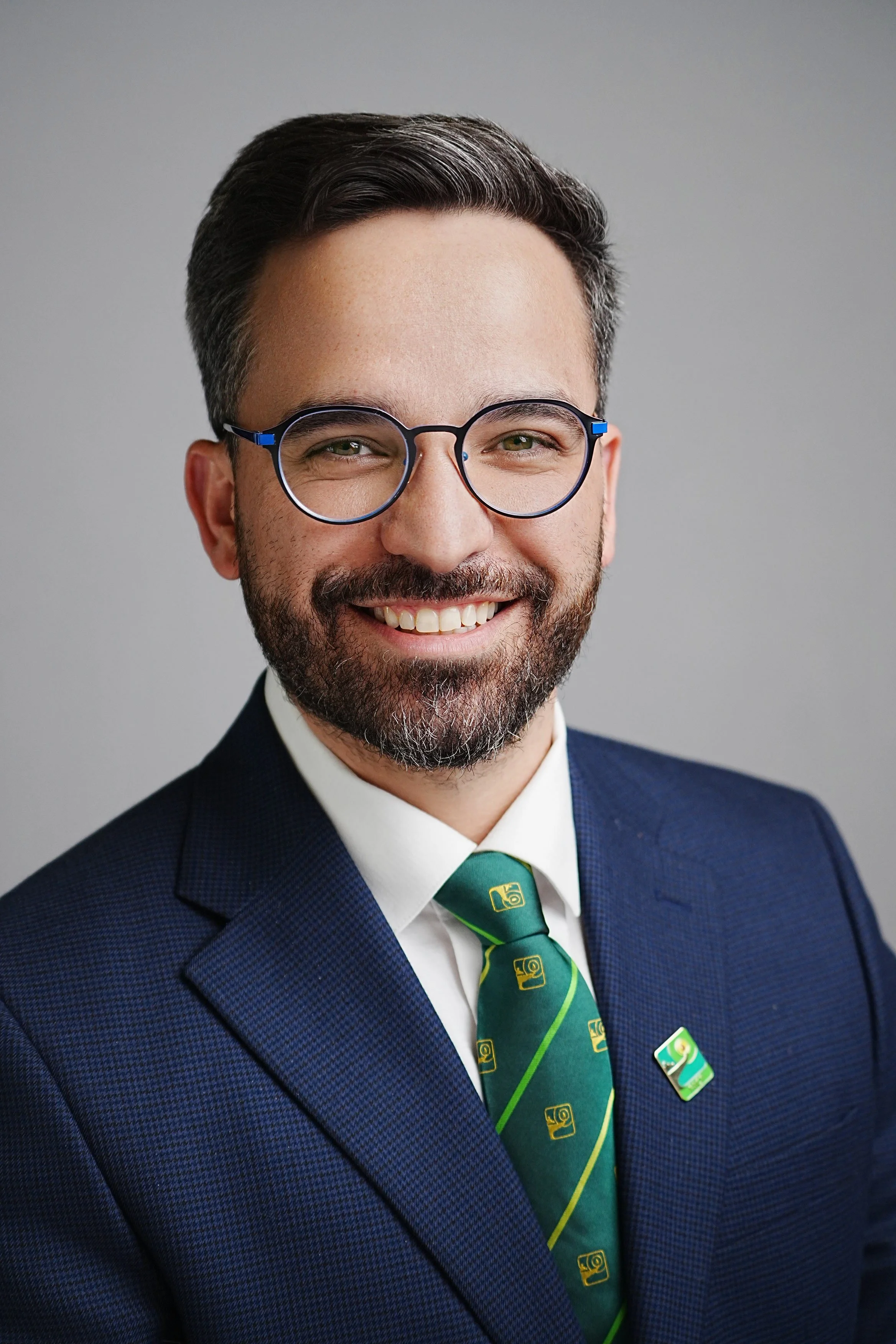 A man with dark hair, glasses, a beard, and wearing a navy blue suit, white shirt, and green tie with a yellow pattern smiling in front of a plain gray background.