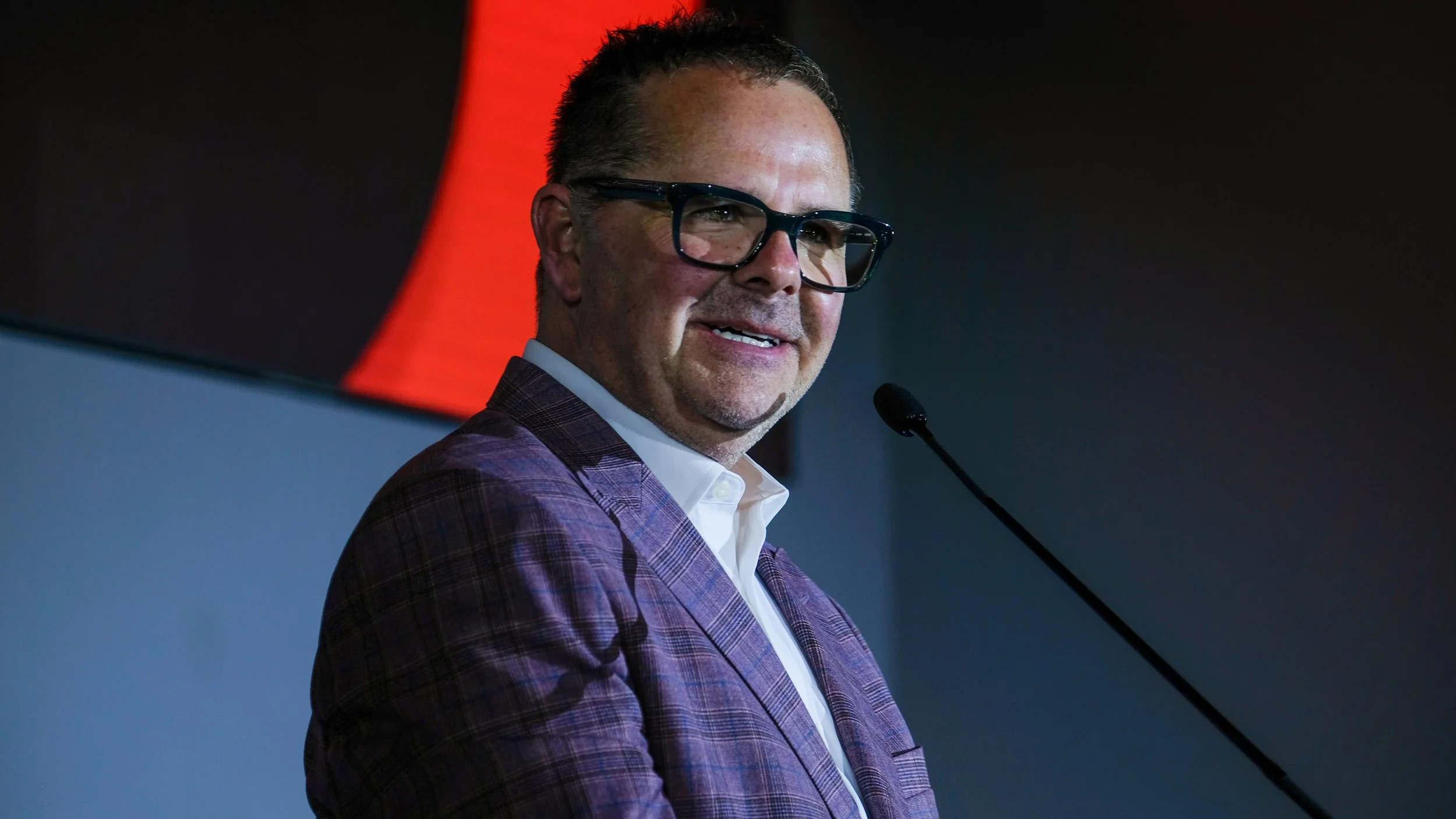A man wearing glasses and a purple checkered blazer speaking at a podium with a microphone, with a red and black background.