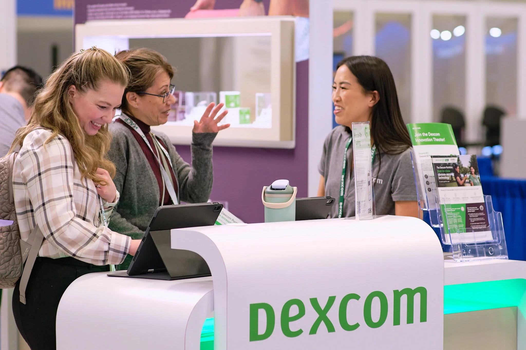Three women are engaged in conversation at a booth with the sign 'pexcom' visible. One woman is wearing glasses and a gray cardigan, another woman has long blonde hair and a checkered shirt, and the third woman is a staff member with long dark hair a