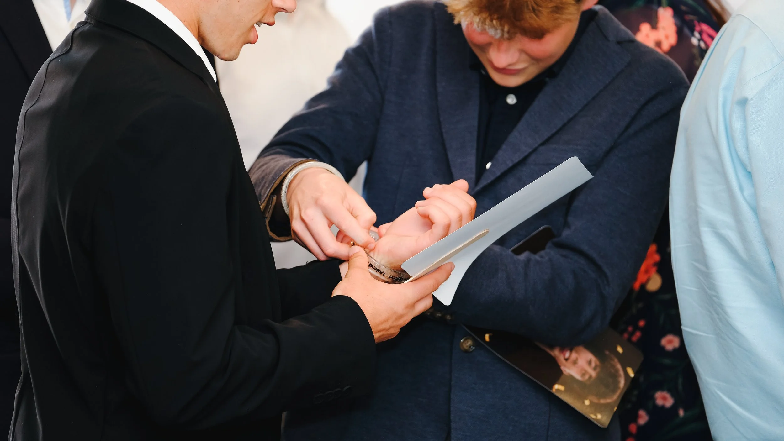 Two men are engaged in an exchange; one is placing a wristband on the other's wrist, while the second man looks down at the wristband. They are at an event or gathering, and the scene is focused on this interaction.