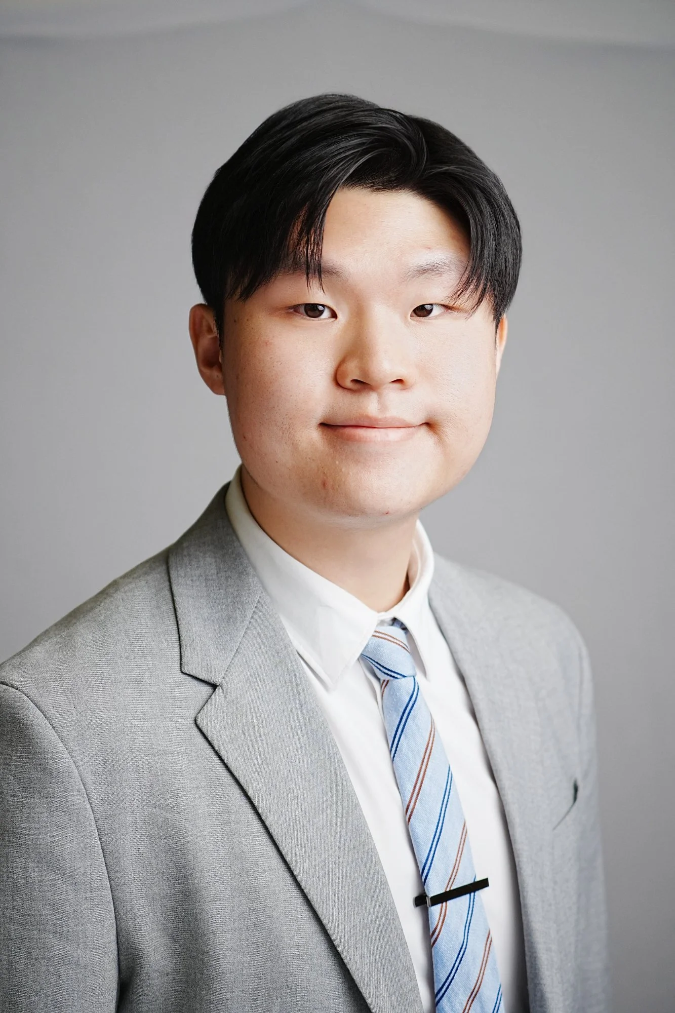 A young man with short black hair wearing a light grey suit, white shirt, and striped tie, posing against a plain grey background.