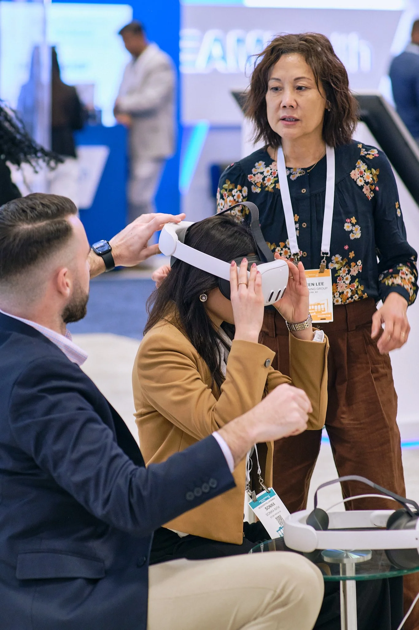 A woman with short brown hair is showing a VR headset to a man in a business suit at a technology convention. Other people are visible in the background at the event.