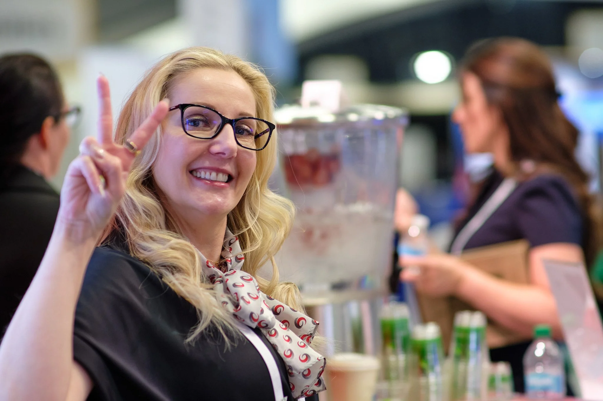 A woman with blonde hair, glasses, and a patterned scarf smiling and making a peace sign at a store or market.