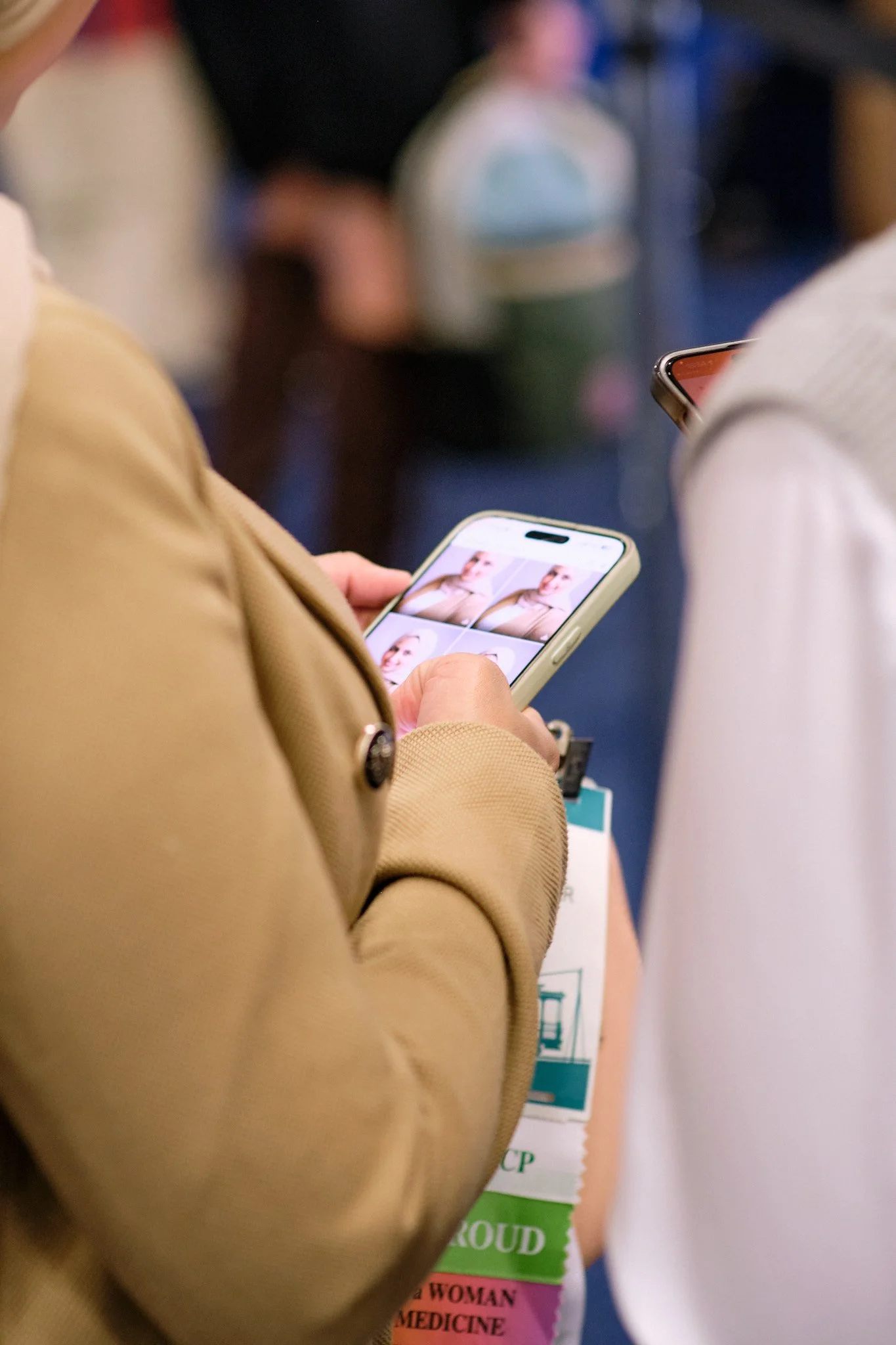 Close-up of a person looking at a smartphone displaying multiple photos of a woman with short hair and a beige blazer. In the background, another person is visible with a badge that reads 'WOMAN IN MEDICINE' in pink.