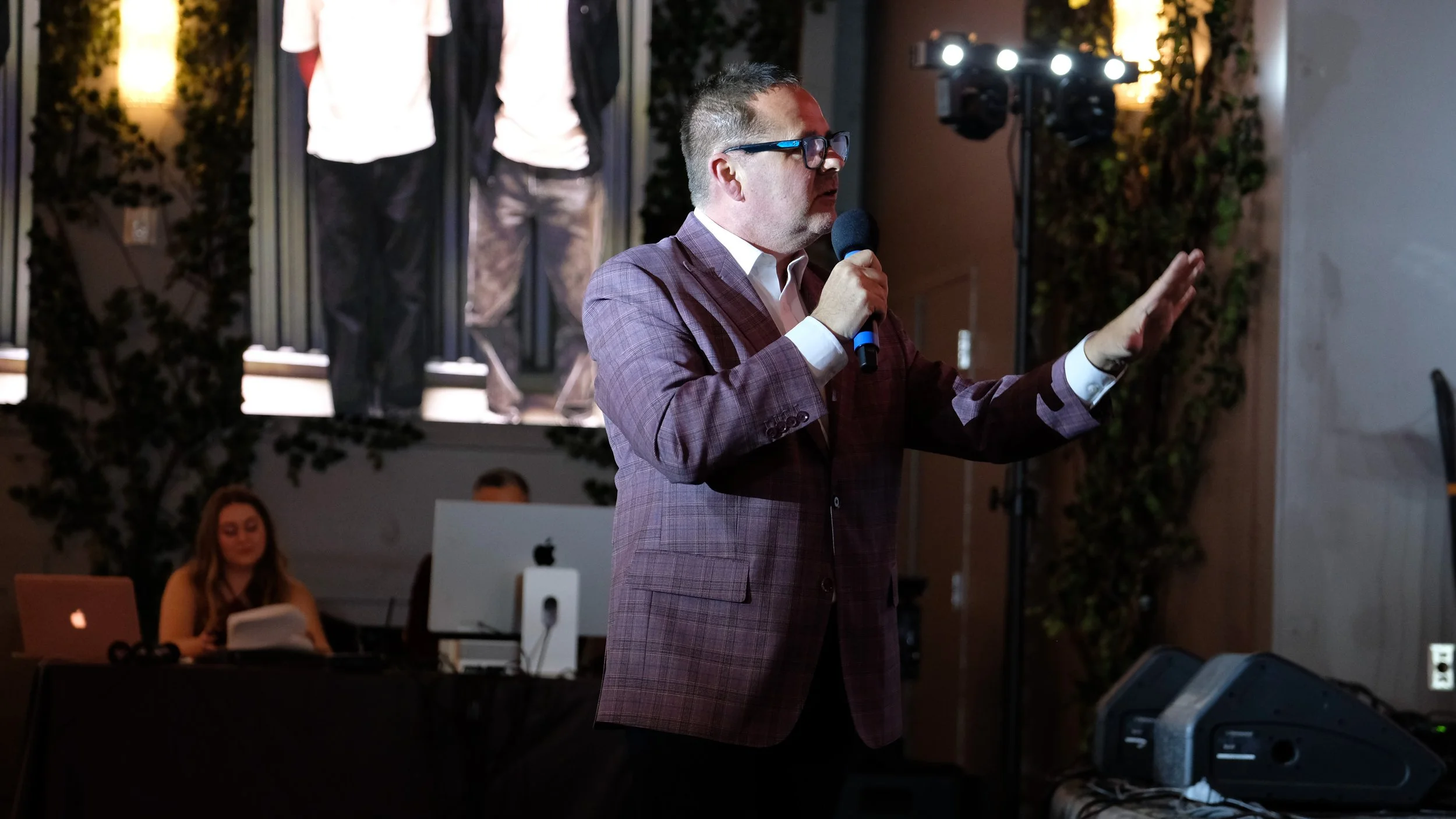Man in a plaid suit and glasses speaking into a microphone at an indoor event, with an audience and a woman working on a laptop in the background.