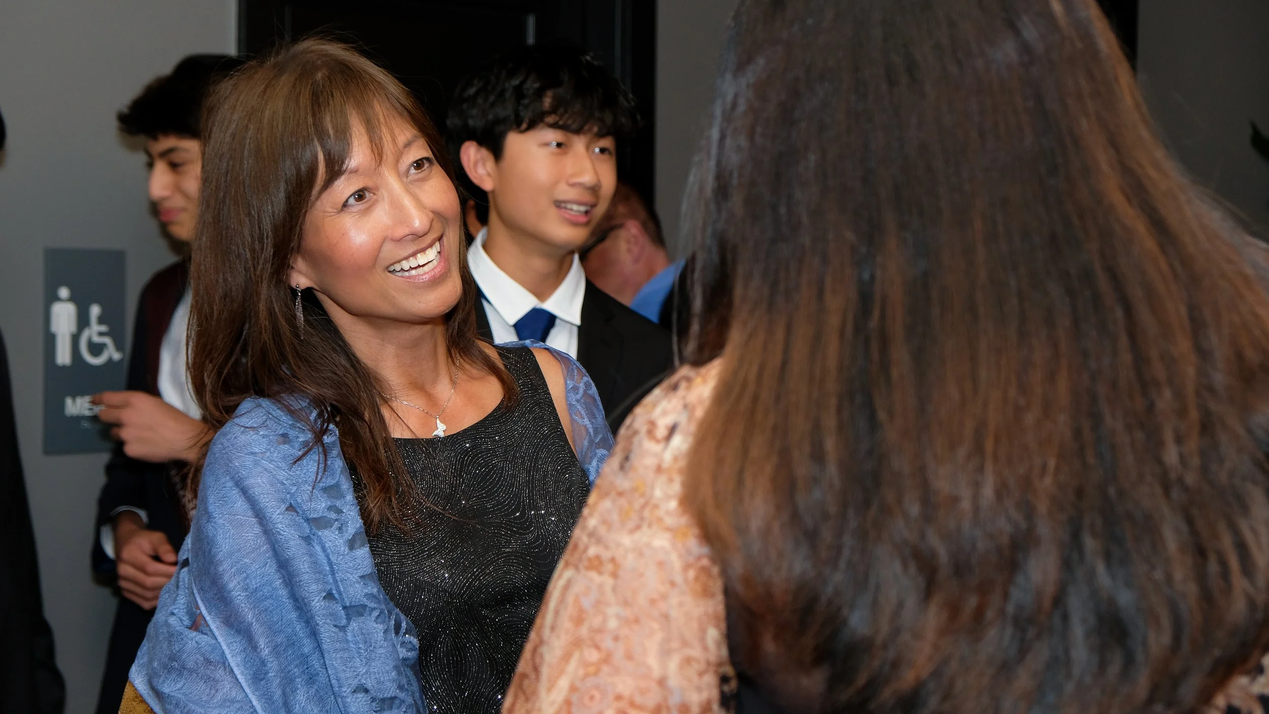 Smiling woman with brown hair wearing a black dress and blue shawl engaged in conversation with a person with long brown hair in a patterned top, at a social event.