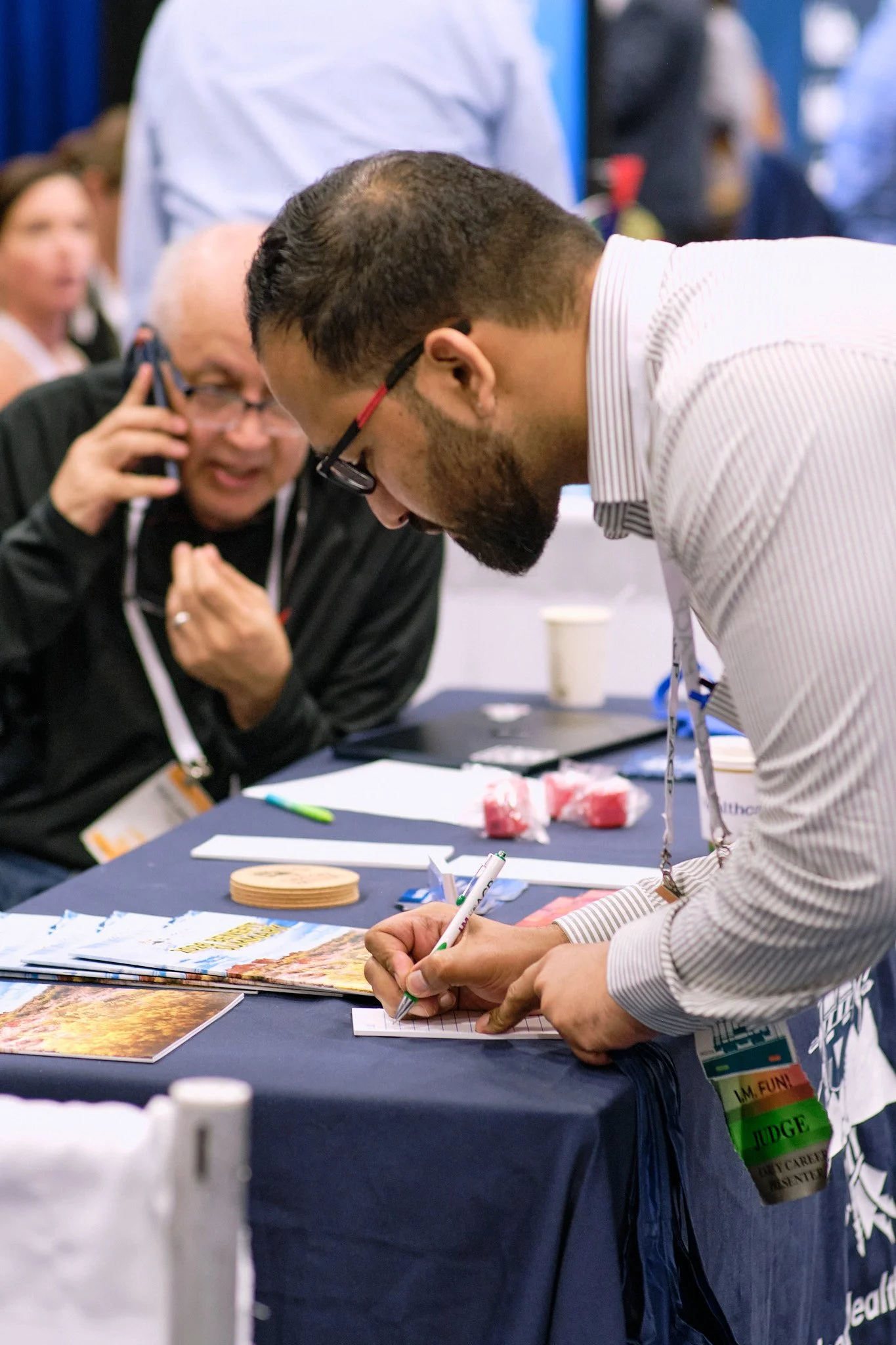 A man with glasses and a beard writing on a notepad at a table with papers and magazines in front of him, at an event or conference.
