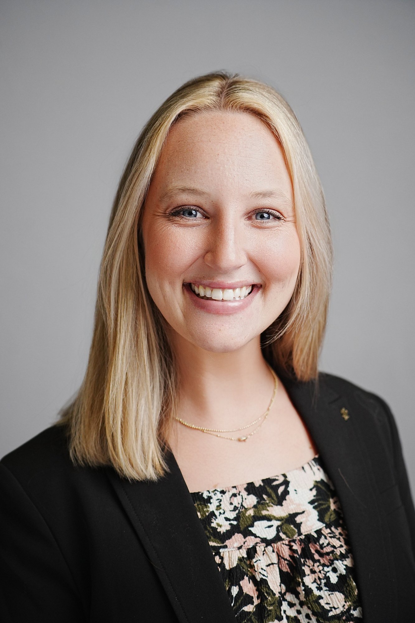 A smiling woman with blonde hair wearing a black blazer and a floral dress, standing against a plain gray background.
