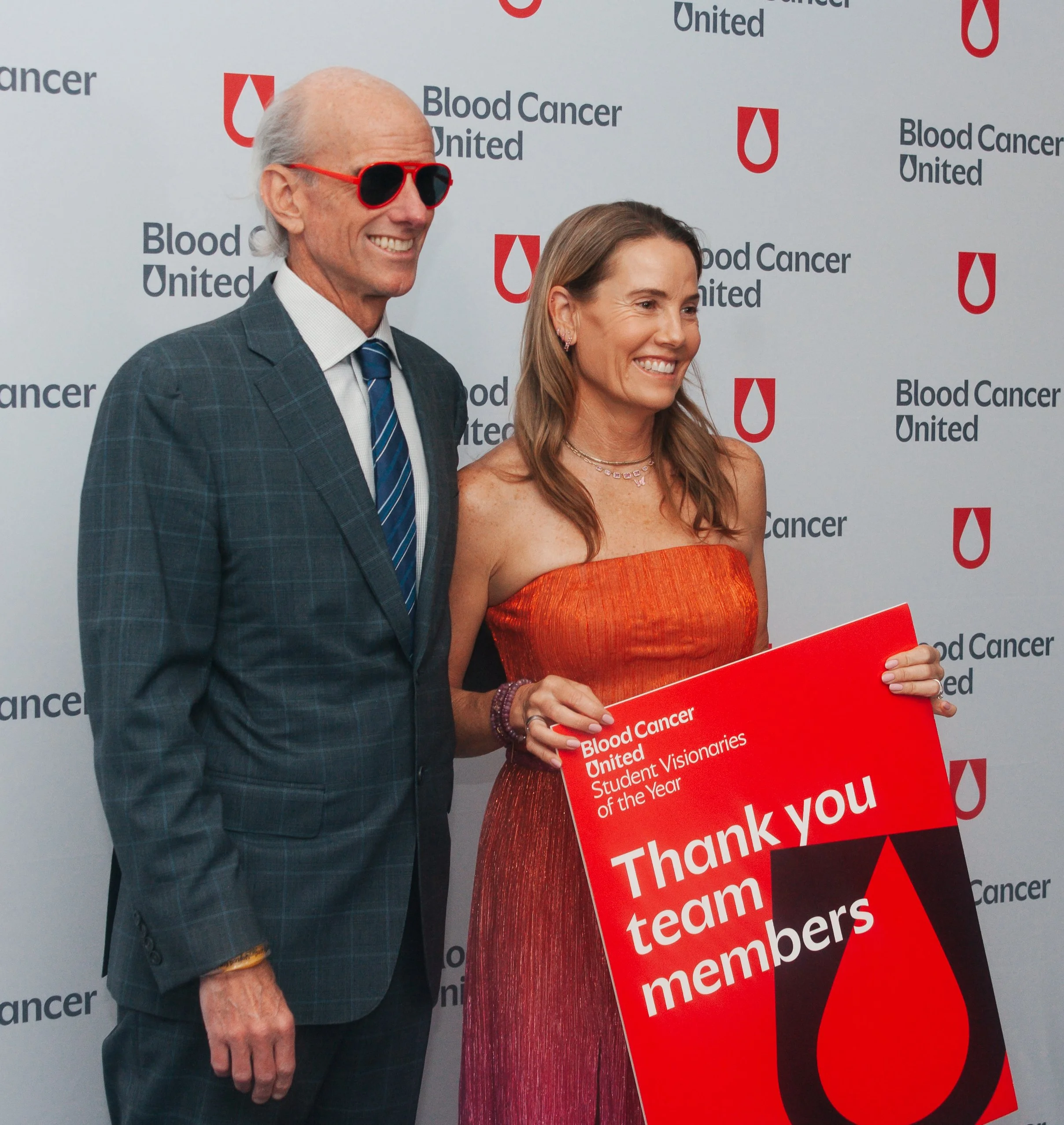 Two smiling individuals, a man in a checkered suit with red sunglasses and a woman in an orange dress, standing in front of a backdrop with the Blood Cancer United logo and text. The woman is holding a red plaque with white text thanking the team and