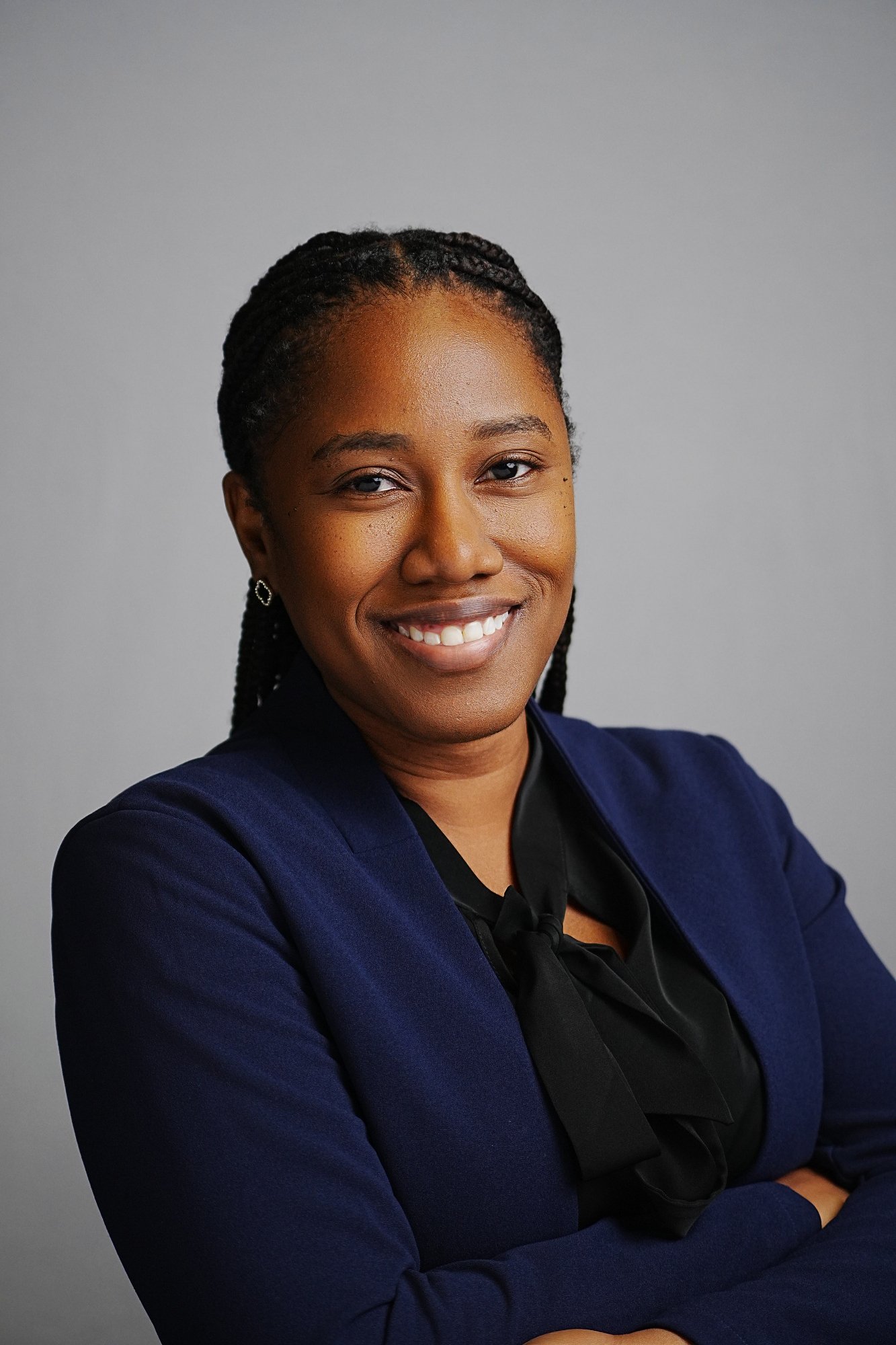 Close-up of a smiling African American woman with braided hair, wearing a dark blue blazer and black top, against a plain gray background.