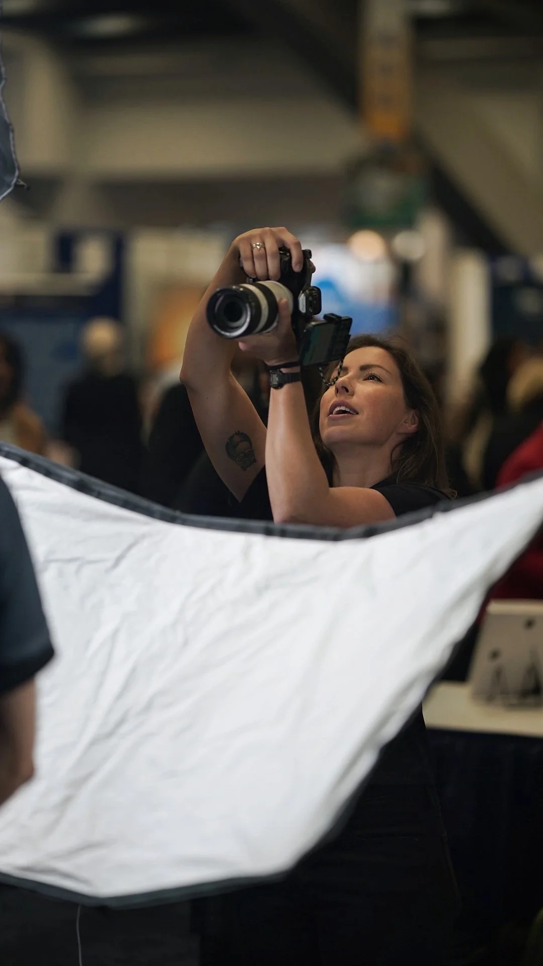 A woman with a tattoo on her arm taking a photograph with a professional camera, looking up in a crowded indoor setting.