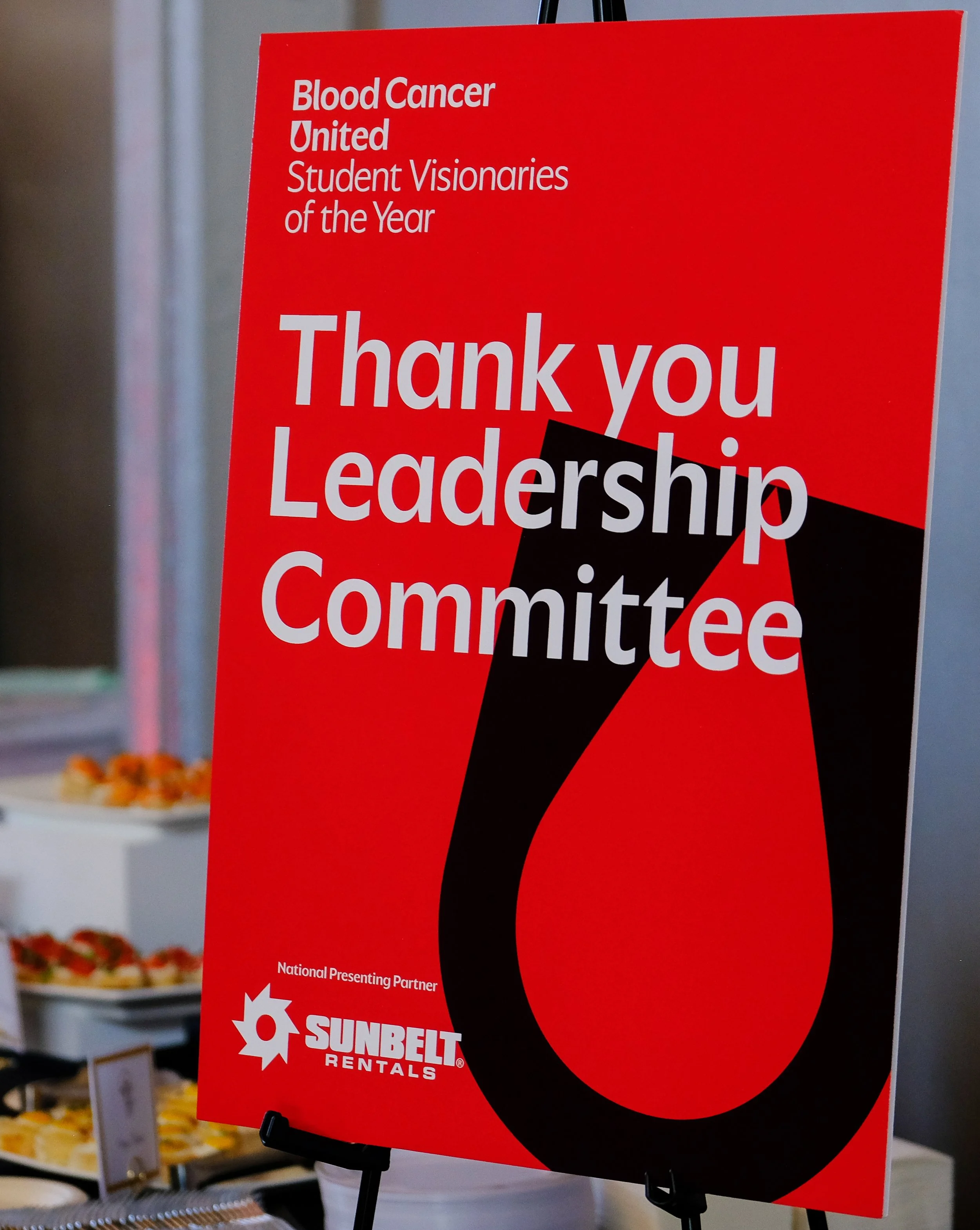 Red sign with white text thanking leadership council, featuring a large black and red number 1 and the Sunbelt Rentals logo at the bottom, located indoors near shelves with food items.