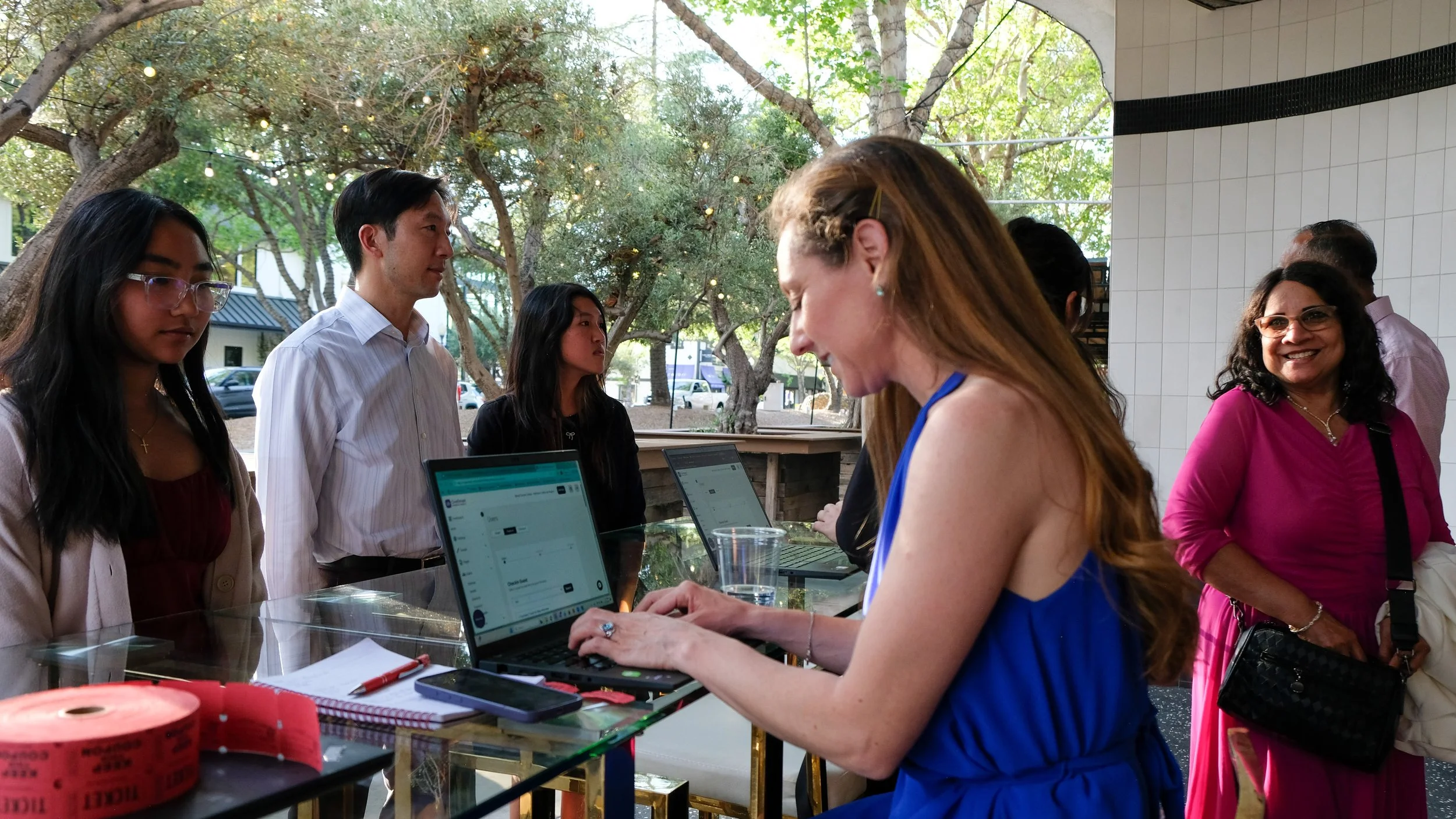 Group of people working on laptops at an outdoor table during daytime, with trees and string lights in the background.