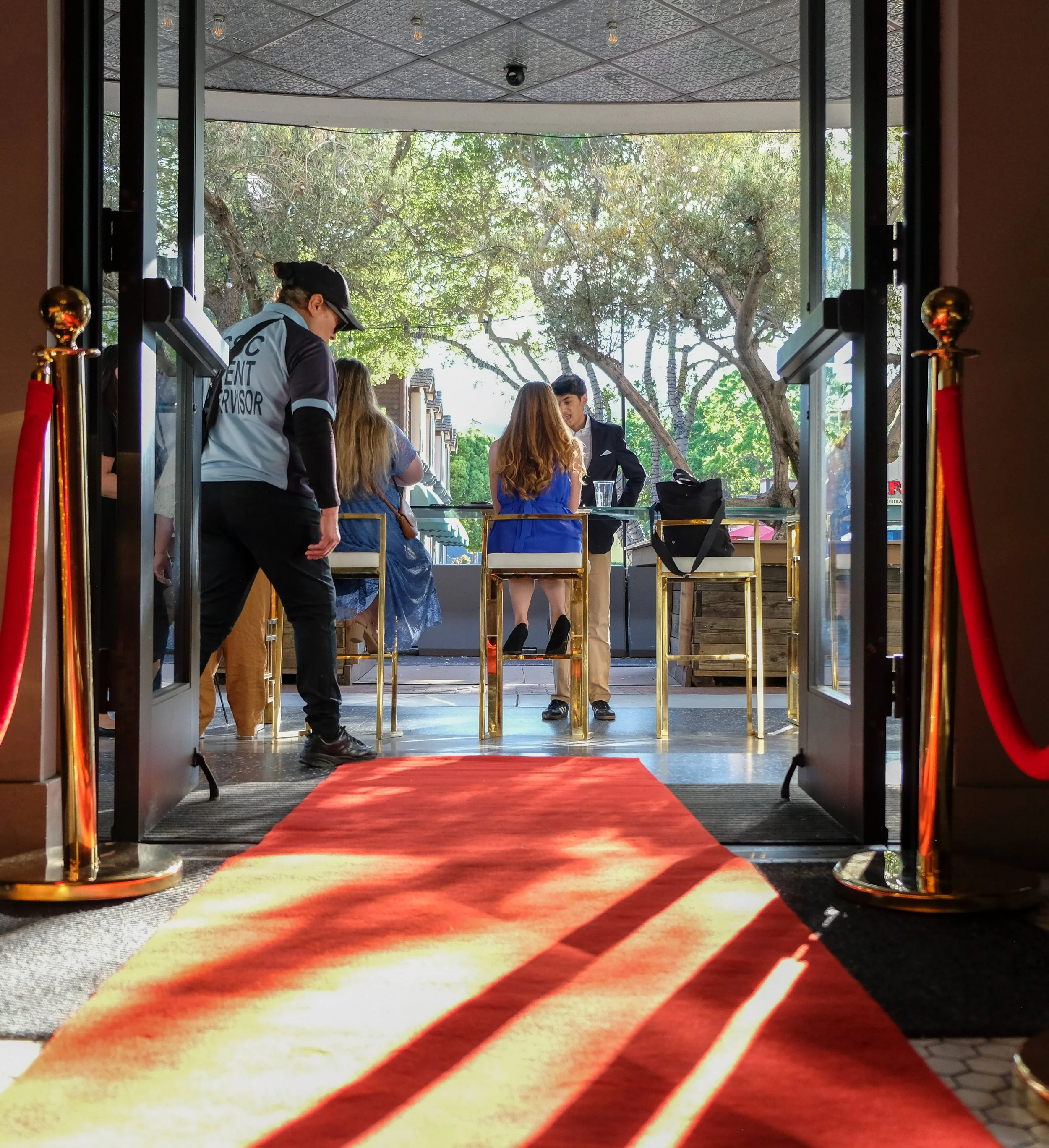 People entering a building through a glass door with gold stanchions and a red carpet, outdoor trees visible in the background.