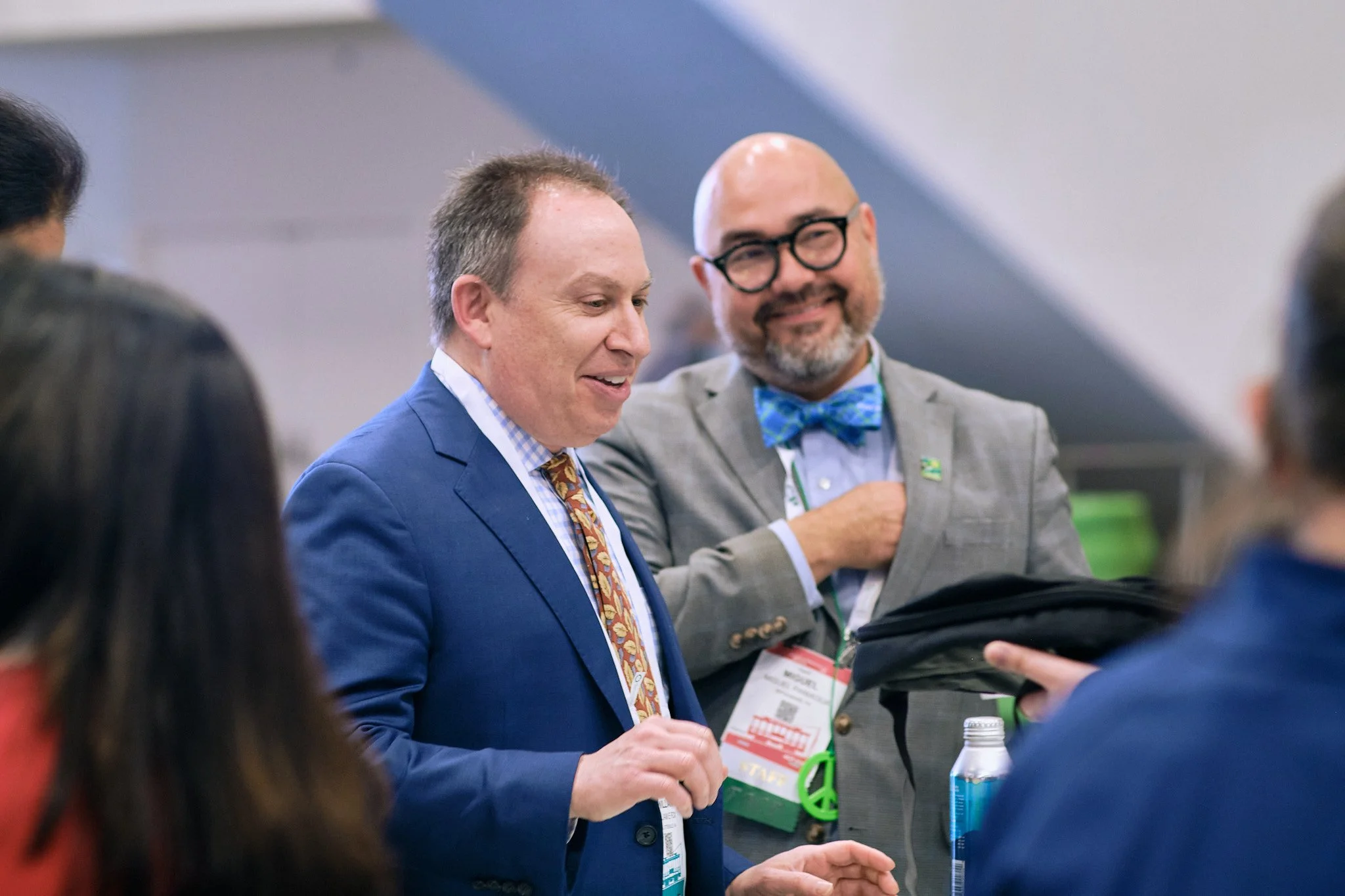 Two men in suits, engaged in conversation, at what appears to be a professional or networking event, with one man smiling and holding a water bottle.