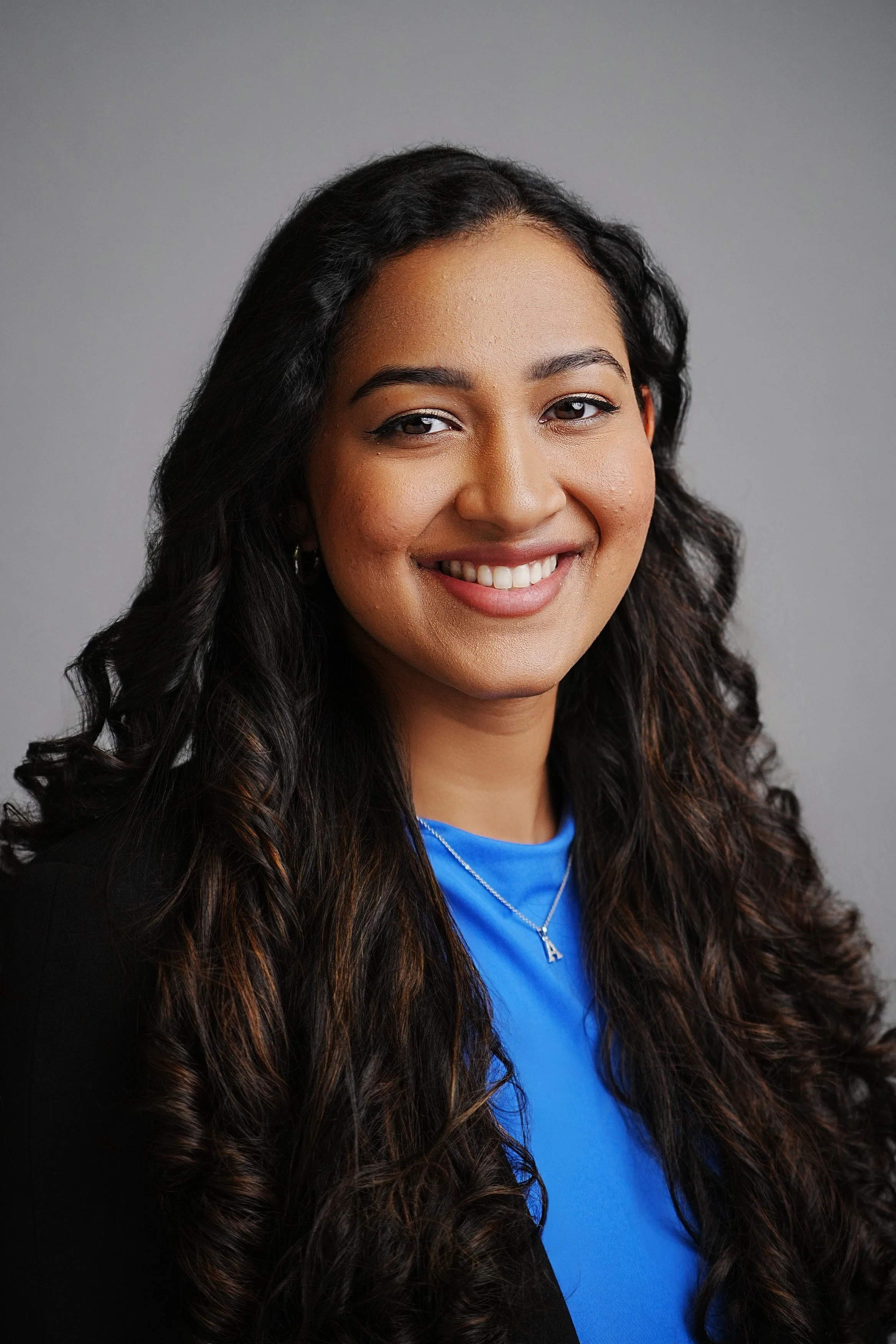 A young woman with long curly dark hair, smiling, wearing a black blazer over a bright blue top, with a silver necklace with an 'A' pendant, against a plain gray background.
