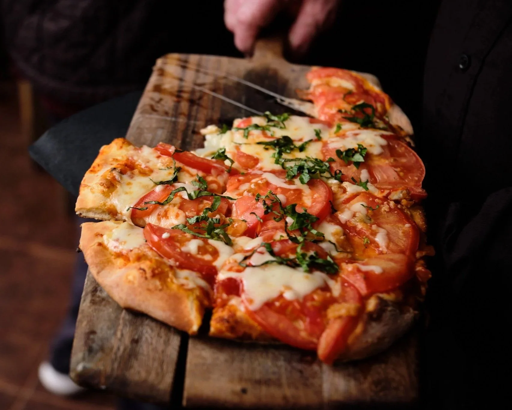 A person holding a wooden pizza peel with a freshly baked pizza topped with tomatoes, cheese, and chopped herbs.