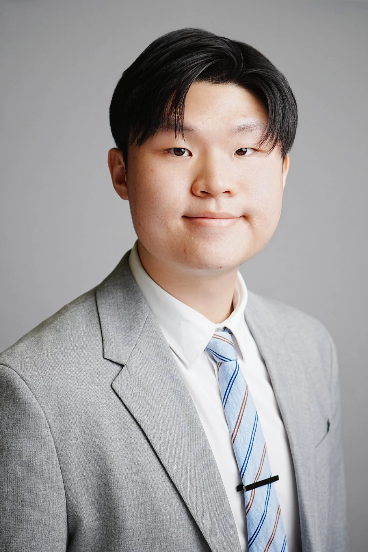 Professional portrait of a young man wearing a light gray suit, white shirt, and striped tie, standing against a plain gray background.