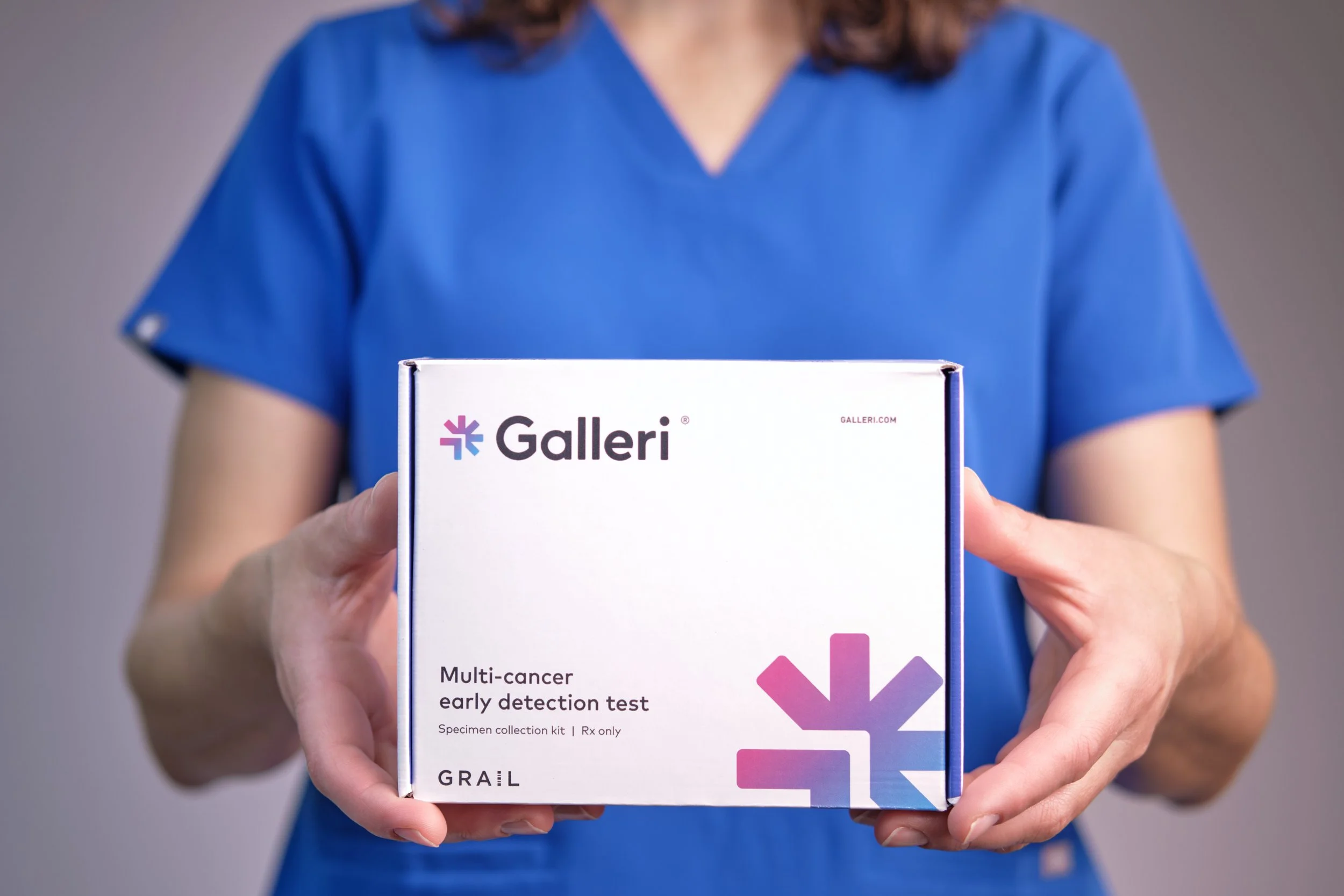 A person in a blue medical uniform holding a clinical test kit box labeled 'Galleri' for multi-cancer early detection, with a plain gray background.