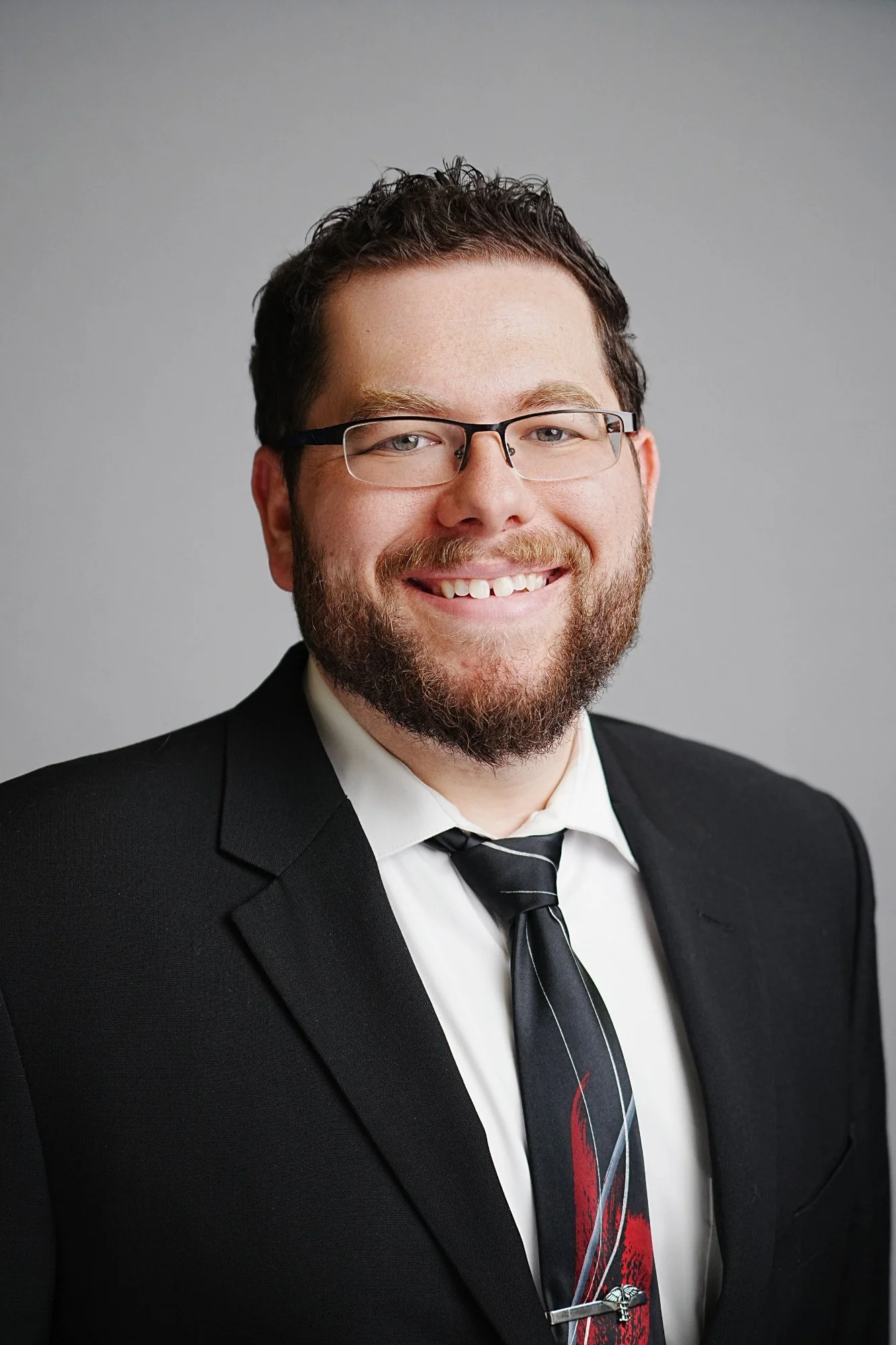 A smiling man with glasses, dark hair, and a beard, dressed in a black suit, white shirt, and holding a black tie, posing against a plain grey background.