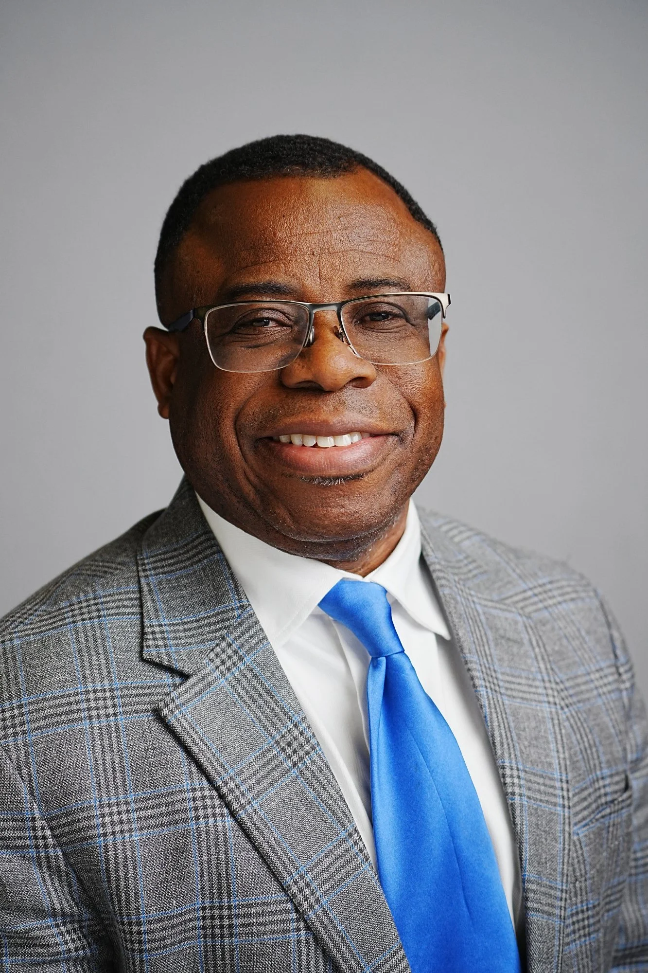 A smiling African American man wearing glasses, a gray plaid suit, white shirt, and bright blue tie, posing against a plain gray background.