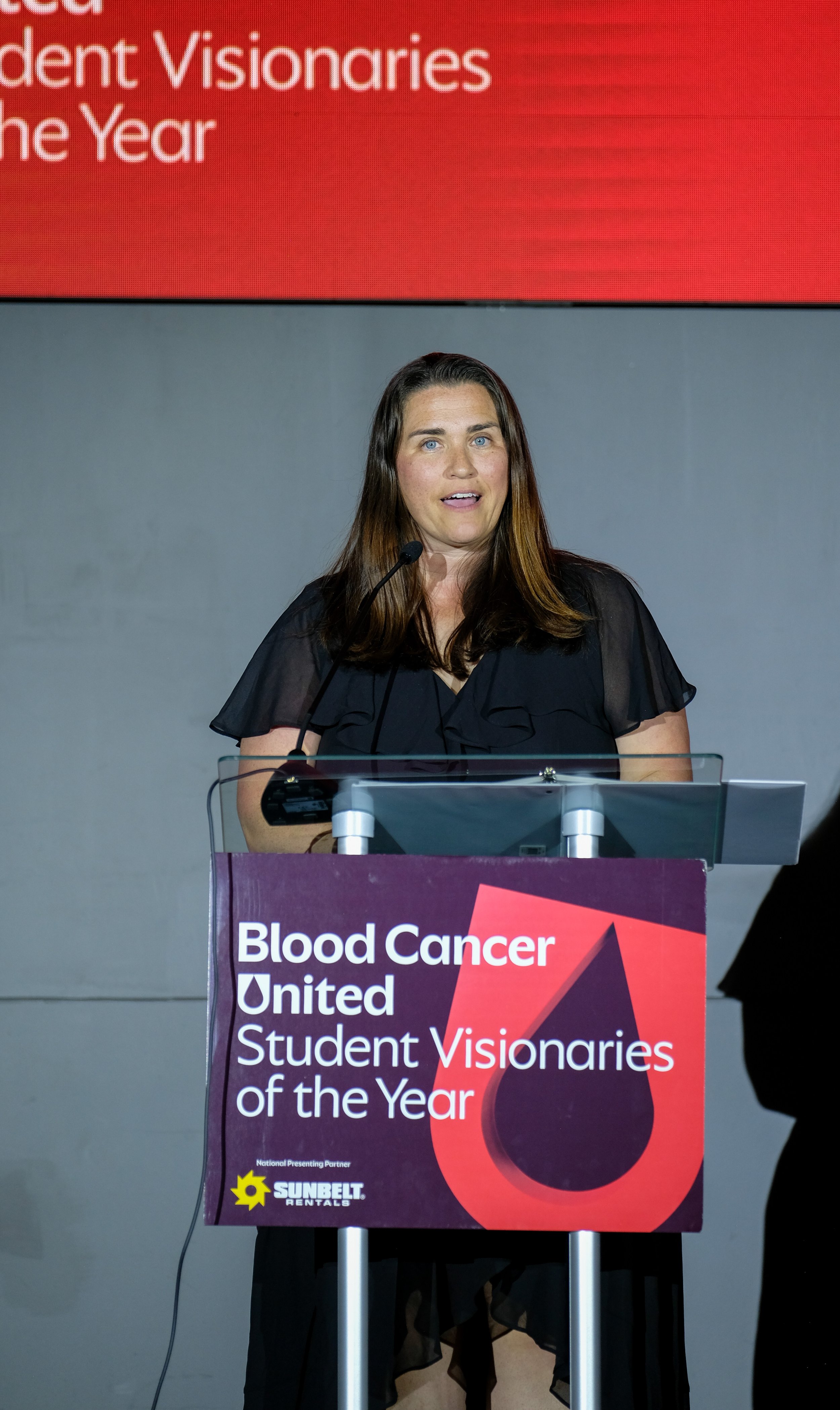 A woman speaking at a podium with a sign that reads "Blood Cancer United Student Visionaries of the Year."
