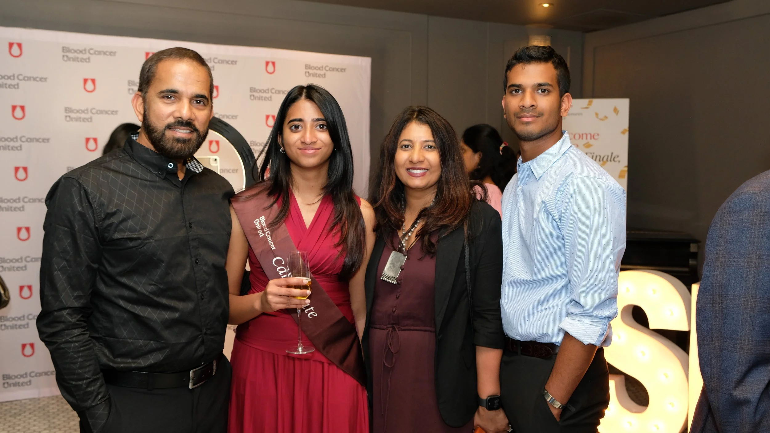 Group of four people smiling at the Blood Cancer United event, with a woman in the center holding a wine glass and wearing a sash.