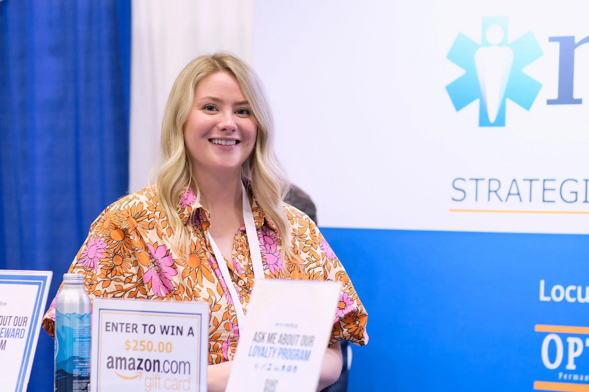 A smiling woman with long blonde hair, wearing a colorful floral dress, standing at a promotional booth with signs for a $250 Amazon gift card contest, at an event with blue and white decorations.