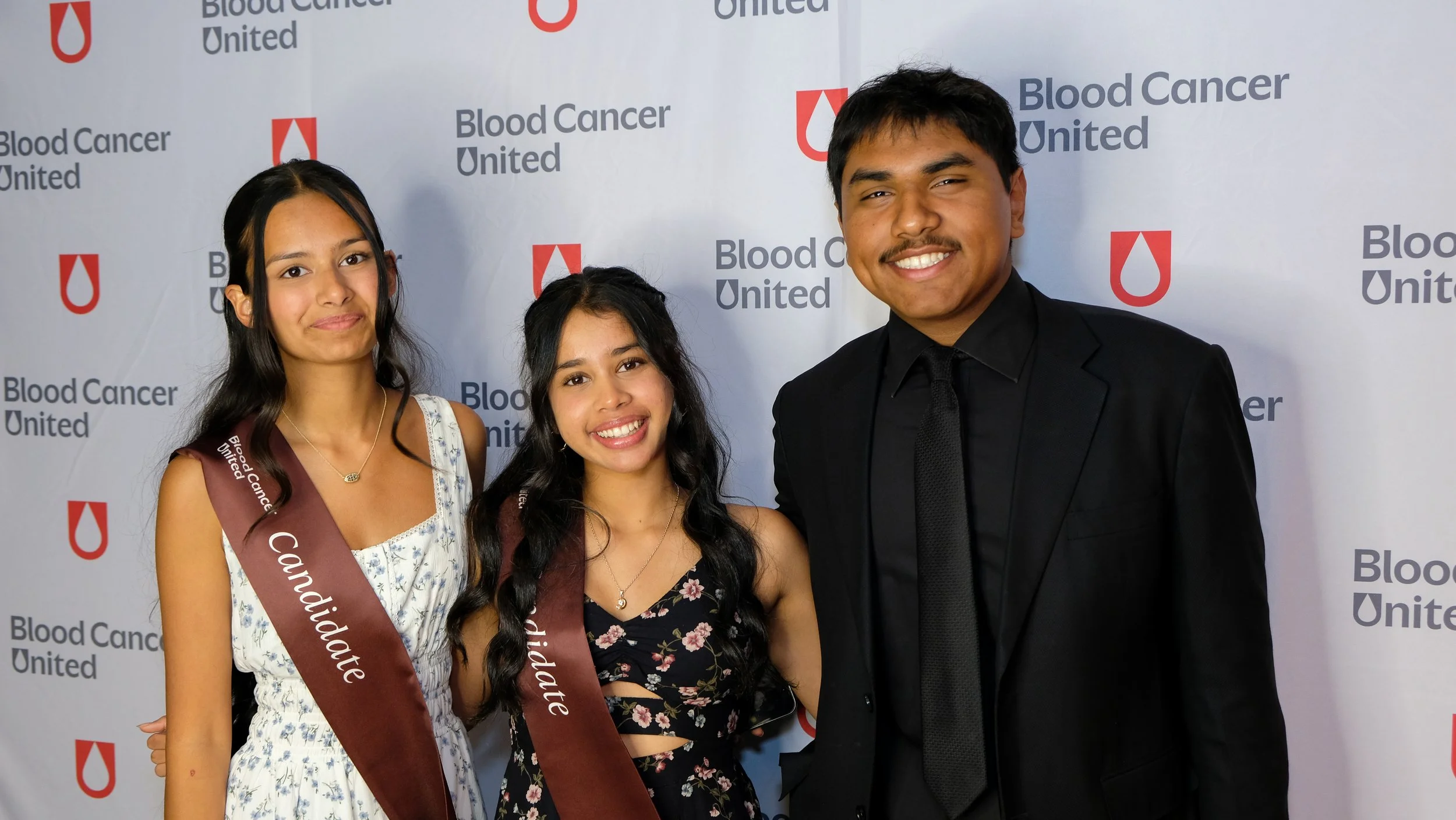 Three young women and one young man standing in front of a backdrop with Blood Cancer United logos. Two women are wearing sashes that say 'Candidate' and 'Congratulations', and they are dressed in formal attire with smiles. The man is wearing a black