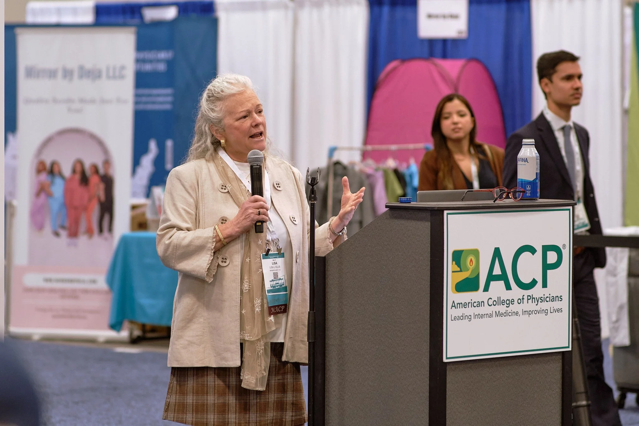 An elderly woman with white hair speaking at a podium with a microphone, wearing a beige jacket with buttons and a lanyard. Behind her, there is a banner for the American College of Physicians (ACP) and two younger people, a woman and a man, listenin