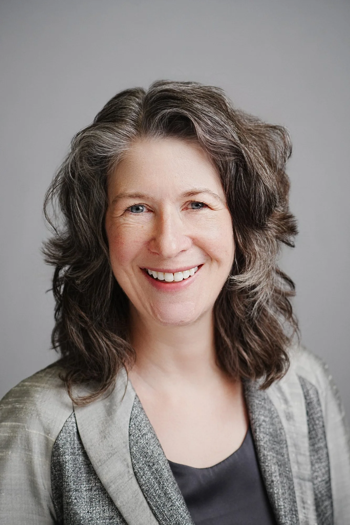 Headshot of a smiling woman with curly gray hair wearing a light blazer over a dark top, against a neutral background.