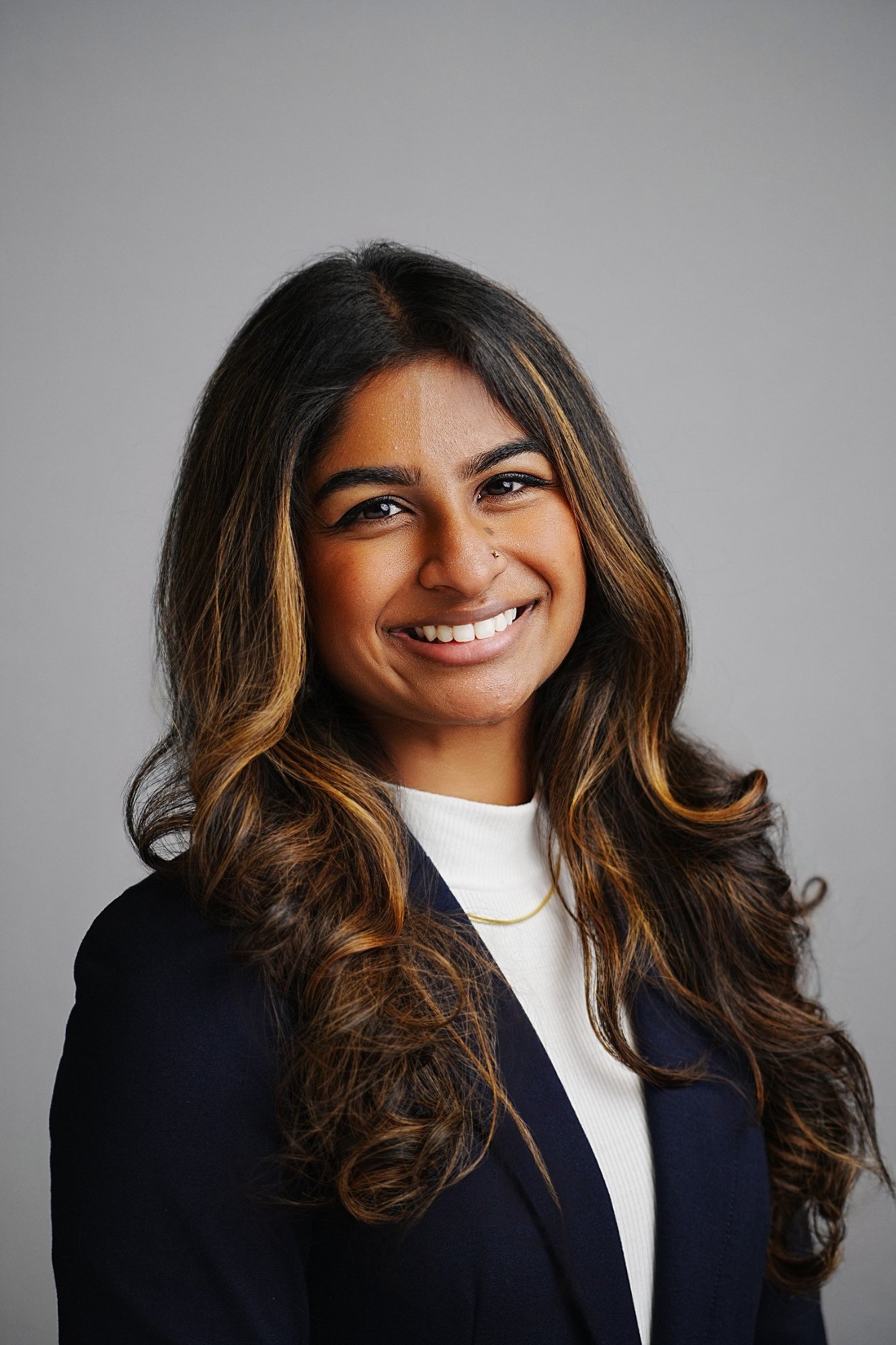 Portrait of a woman with long, wavy brown hair with blonde highlights, smiling, wearing a black blazer over a white top, with a gray background.