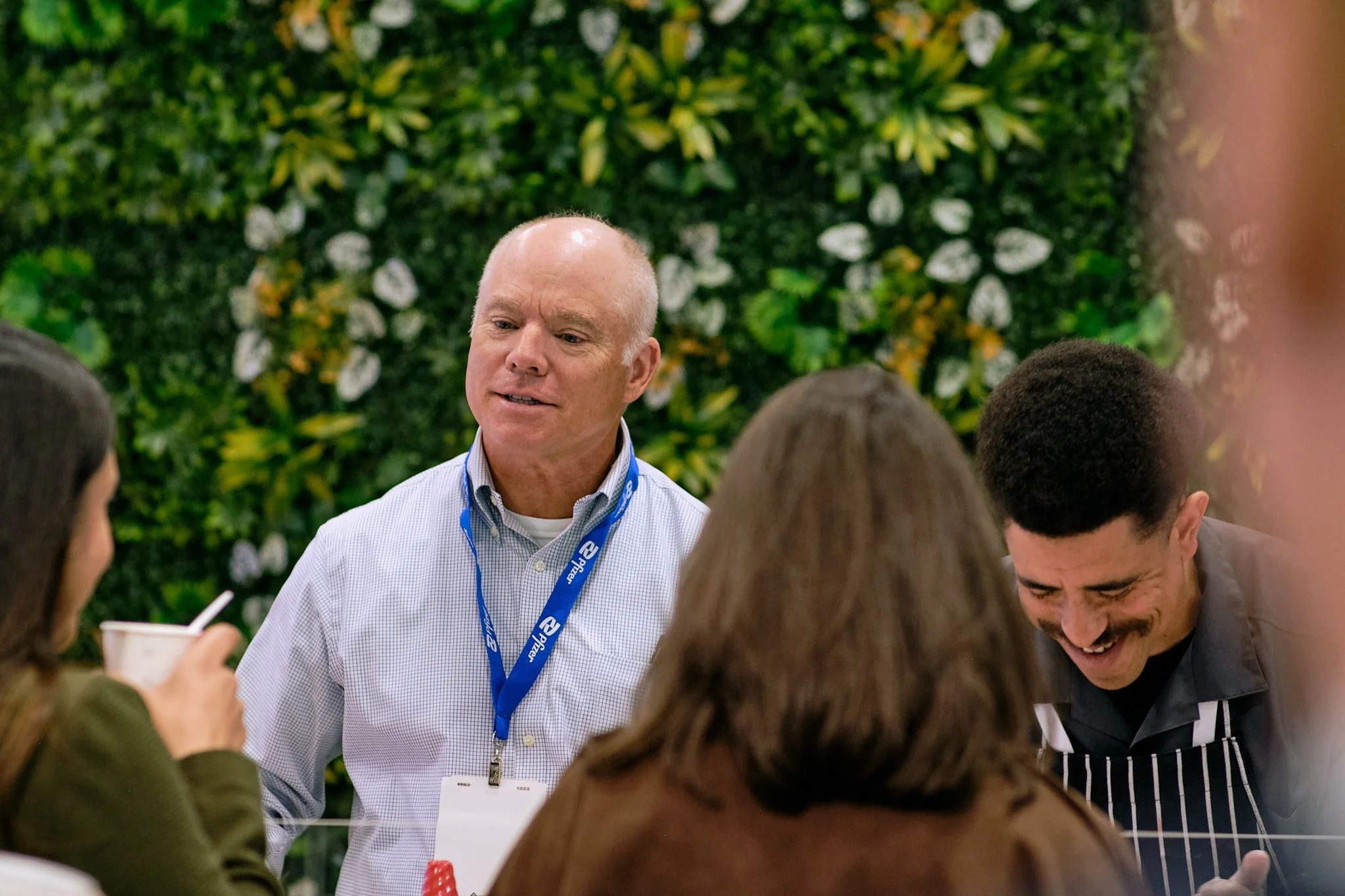 A group of diverse young people engaging in conversation with an older man at a gathering with a lush green background.